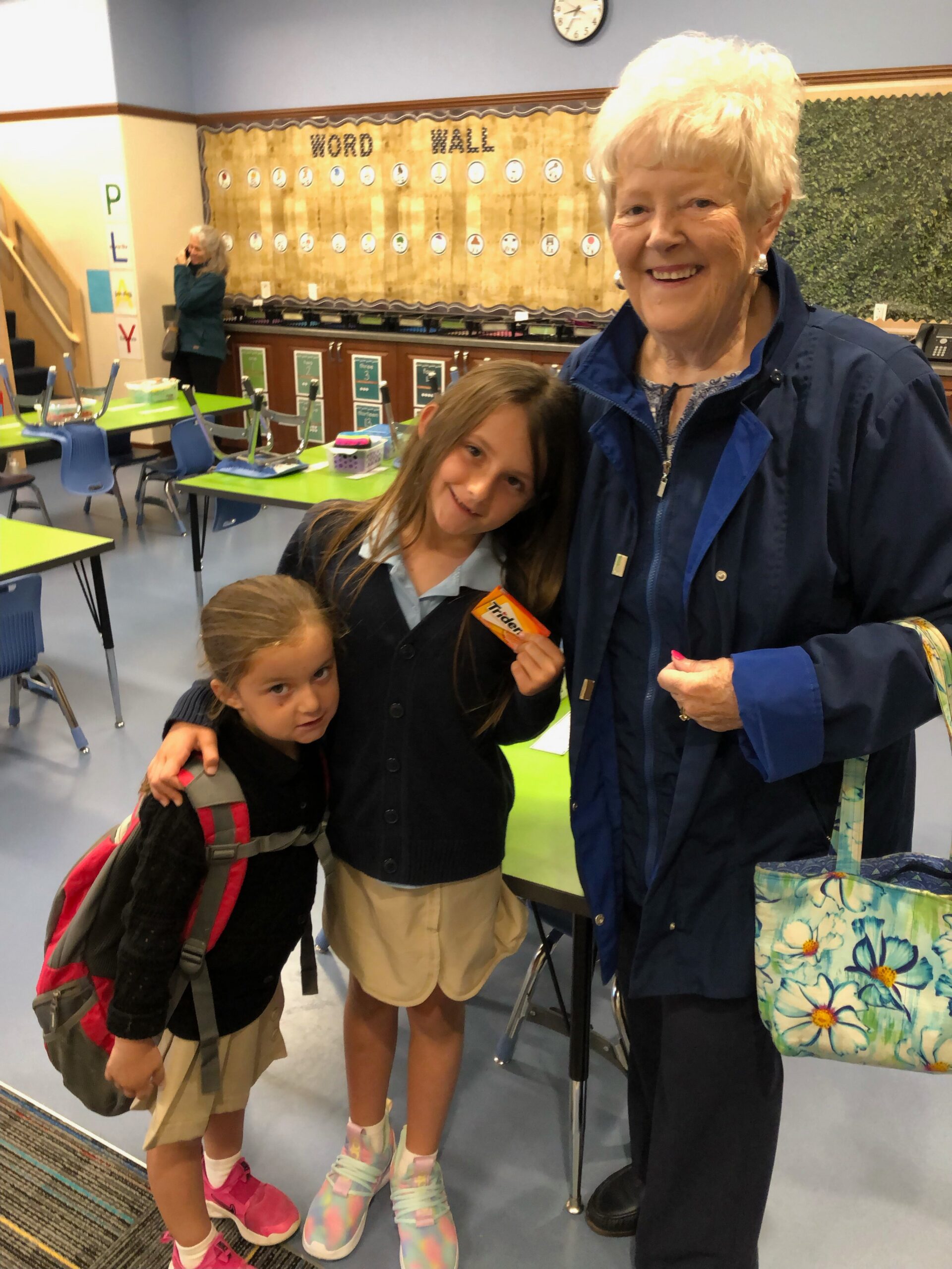 Smiling older woman with two children in a classroom, word wall behind them and backpacks on tables.