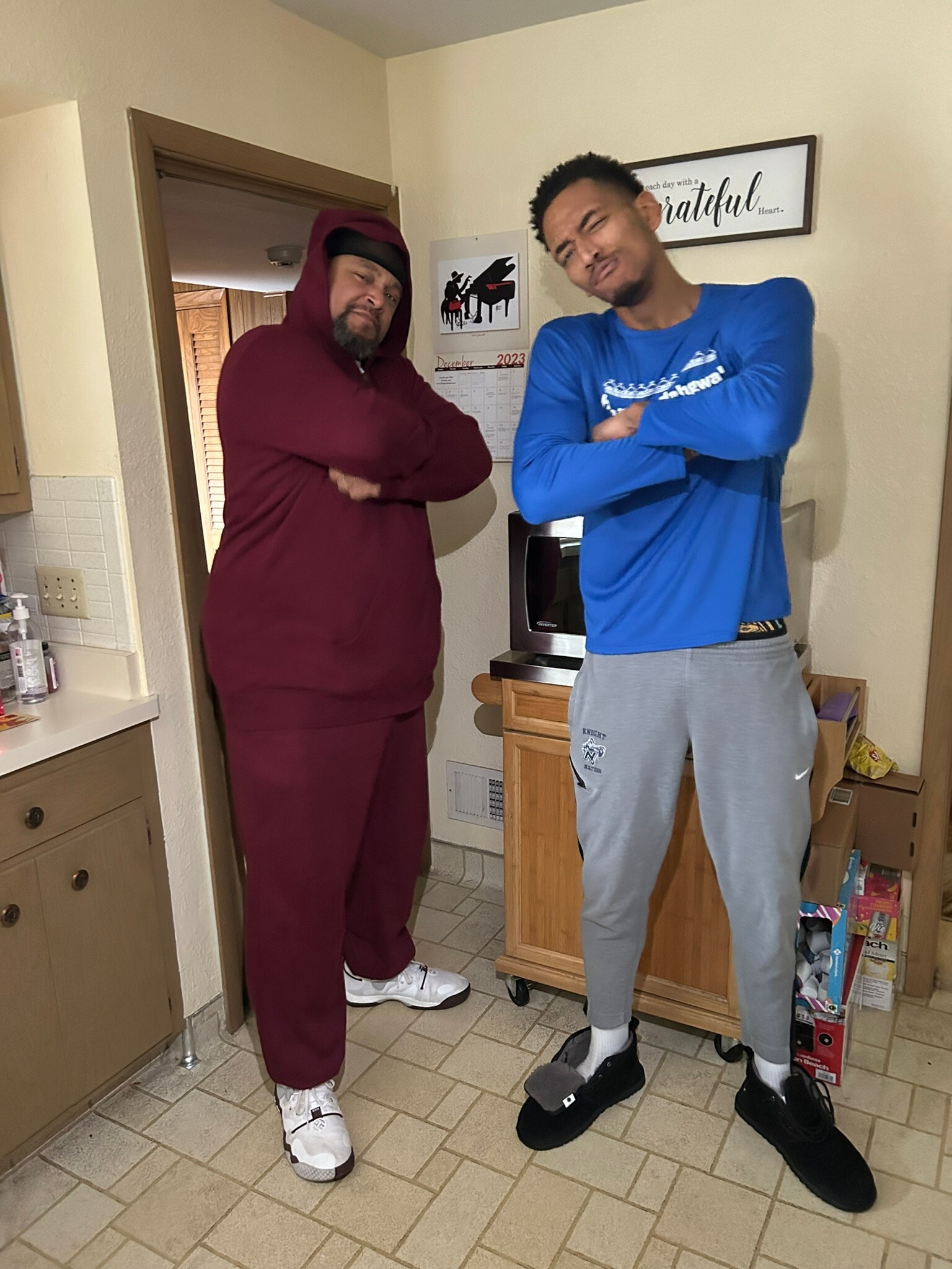 Two men pose with arms crossed in a home kitchen, one in maroon sweats and white sneakers, the other in a blue long-sleeve shirt and gray sweatpants with black slides.