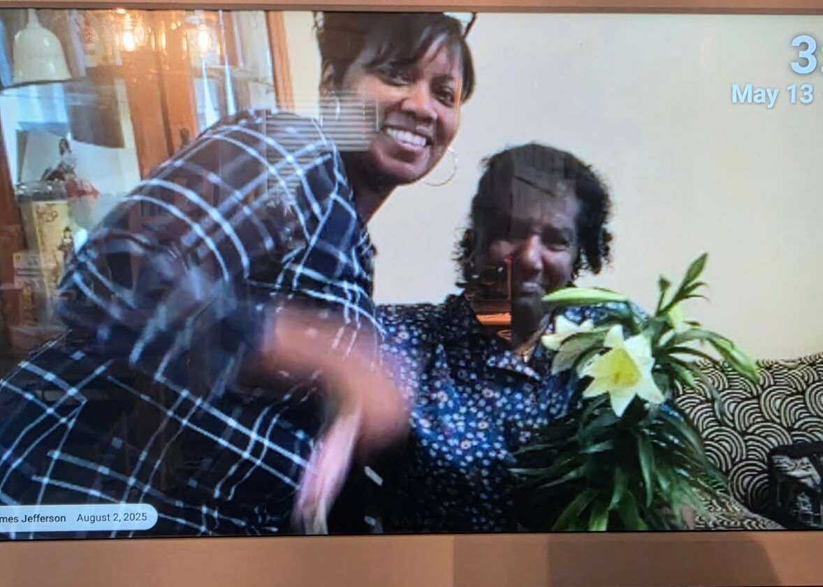 Two women smiling during a video call, one standing and the other seated with a bouquet of lilies on a couch in the background