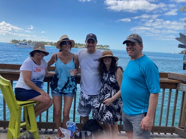 Five friends posing together on a sunlit pier with turquoise water and boats in the background.