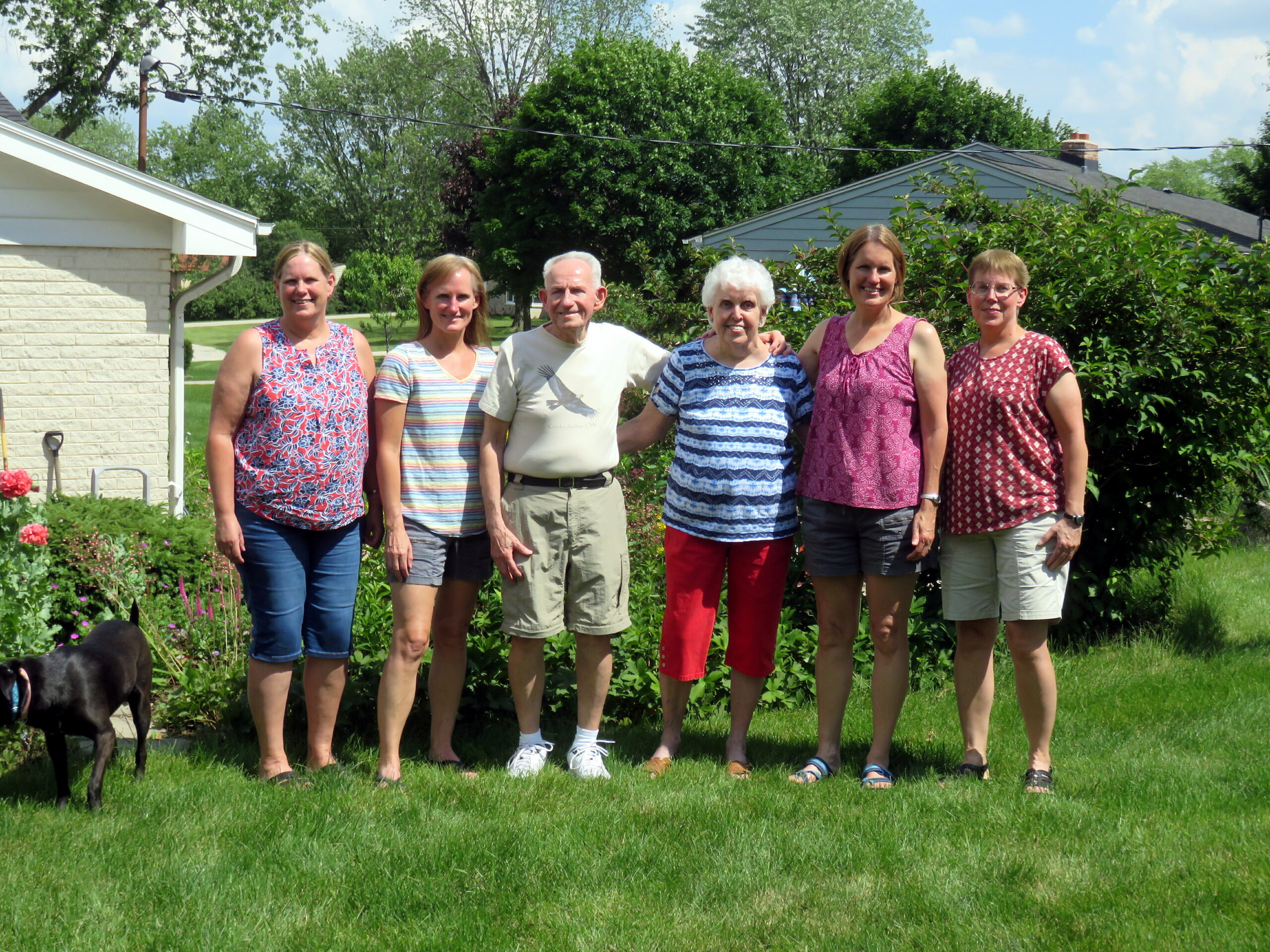 Six adults stand together in a sunny backyard garden with green plants and trees in the background; a black dog is at the left edge.