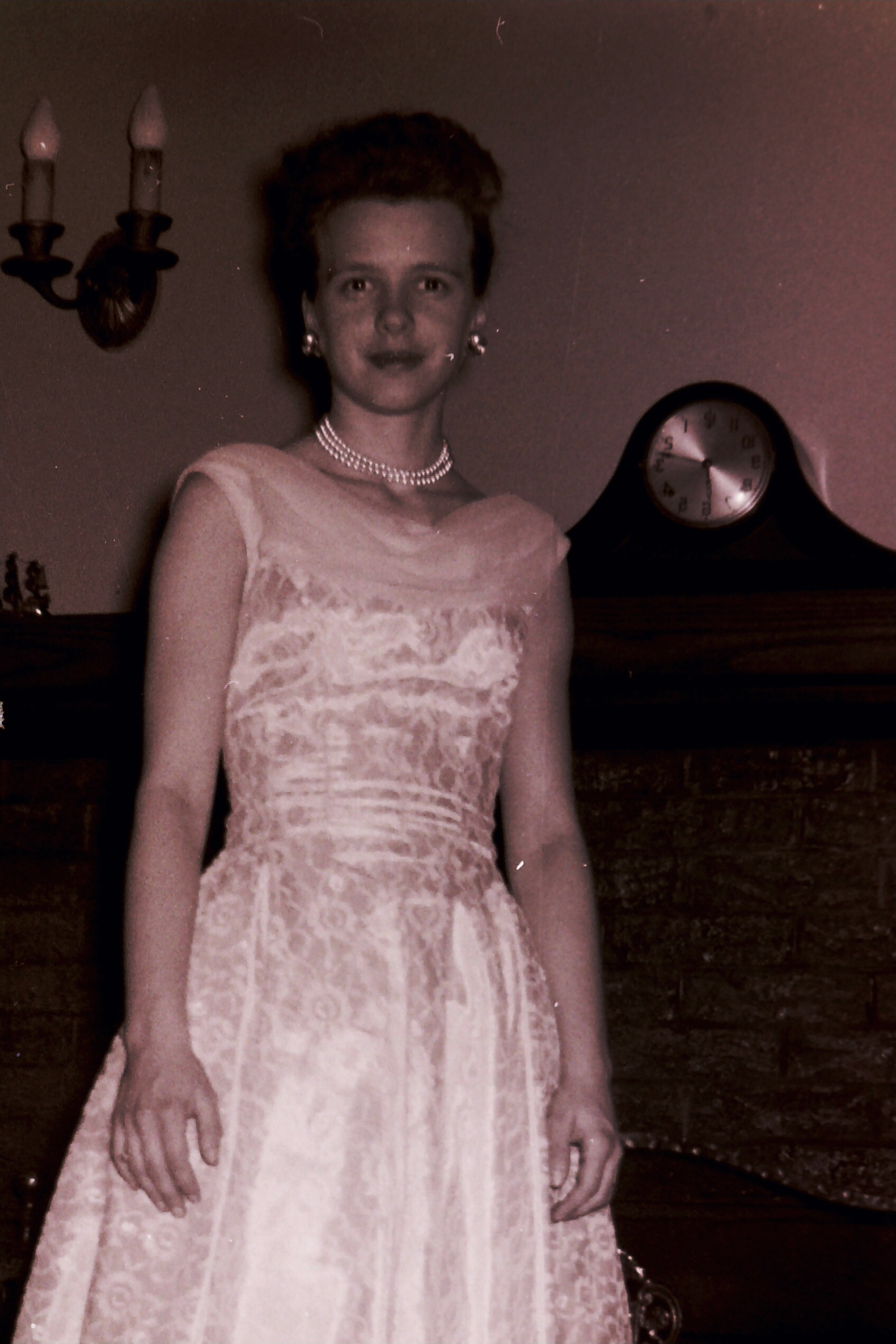 Woman in a lace dress with pearls posing indoors beside a mantel clock behind her.