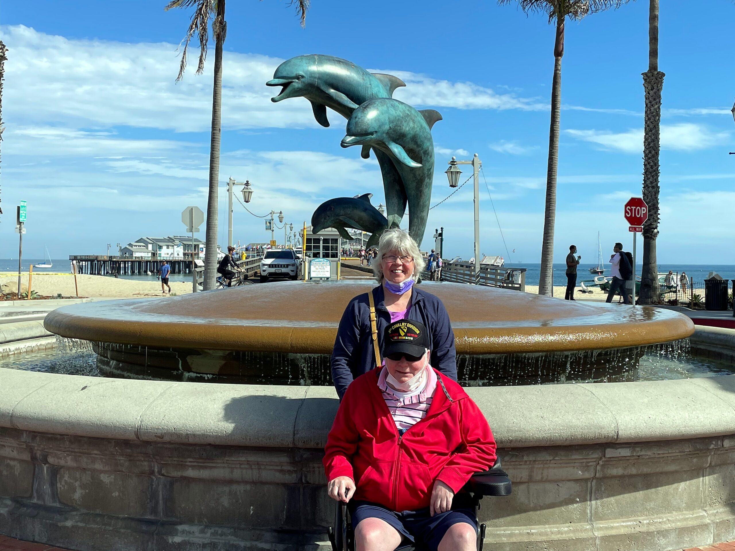 Two people in a wheelchair pose in front of a circular fountain with teal dolphin sculptures on a sunny seaside boardwalk, palm trees behind them.