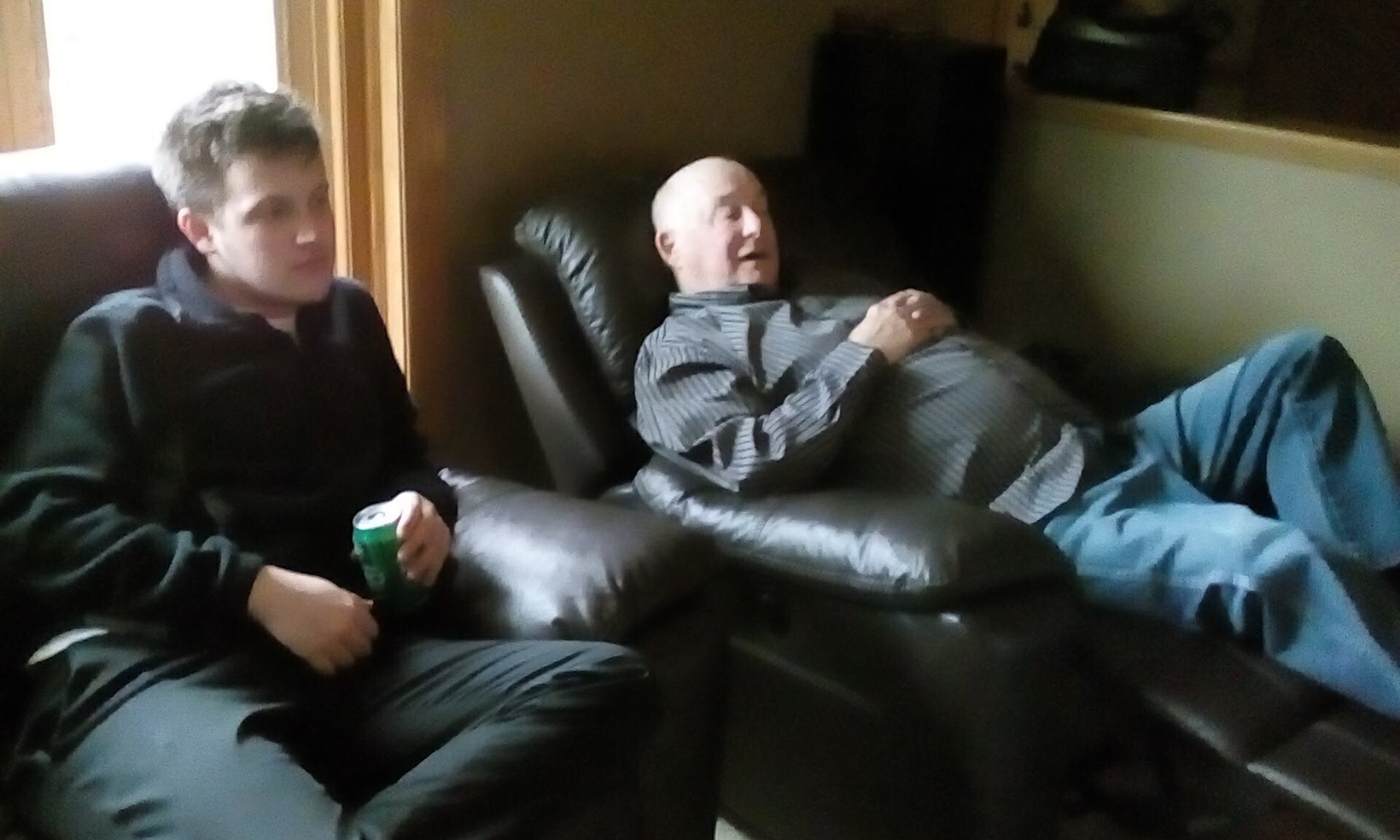 Two men sit on dark leather couches in a living room; the younger man on the left holds a green can while the older man reclines and smiles in the center.