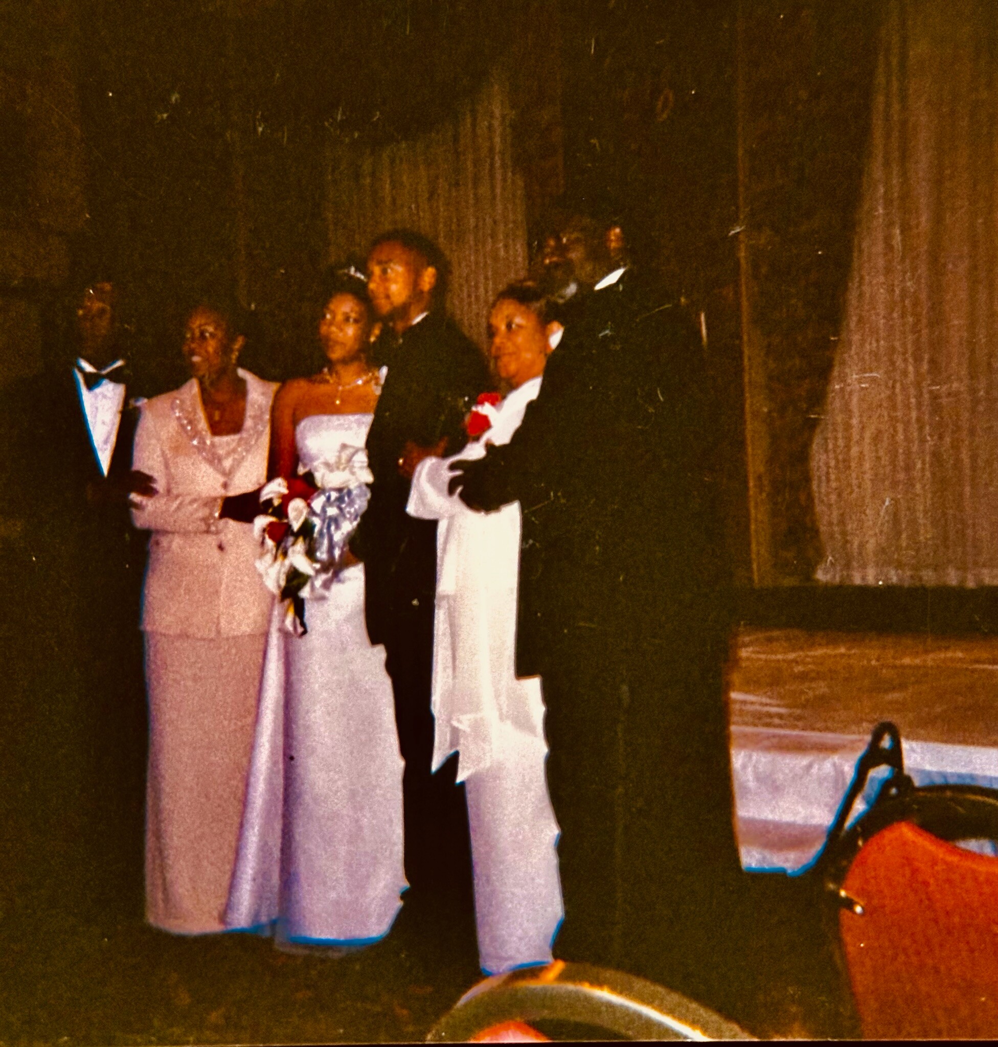 Group of people in formal attire posing on a stage; two women in light dresses hold bouquets, others in dark suits.