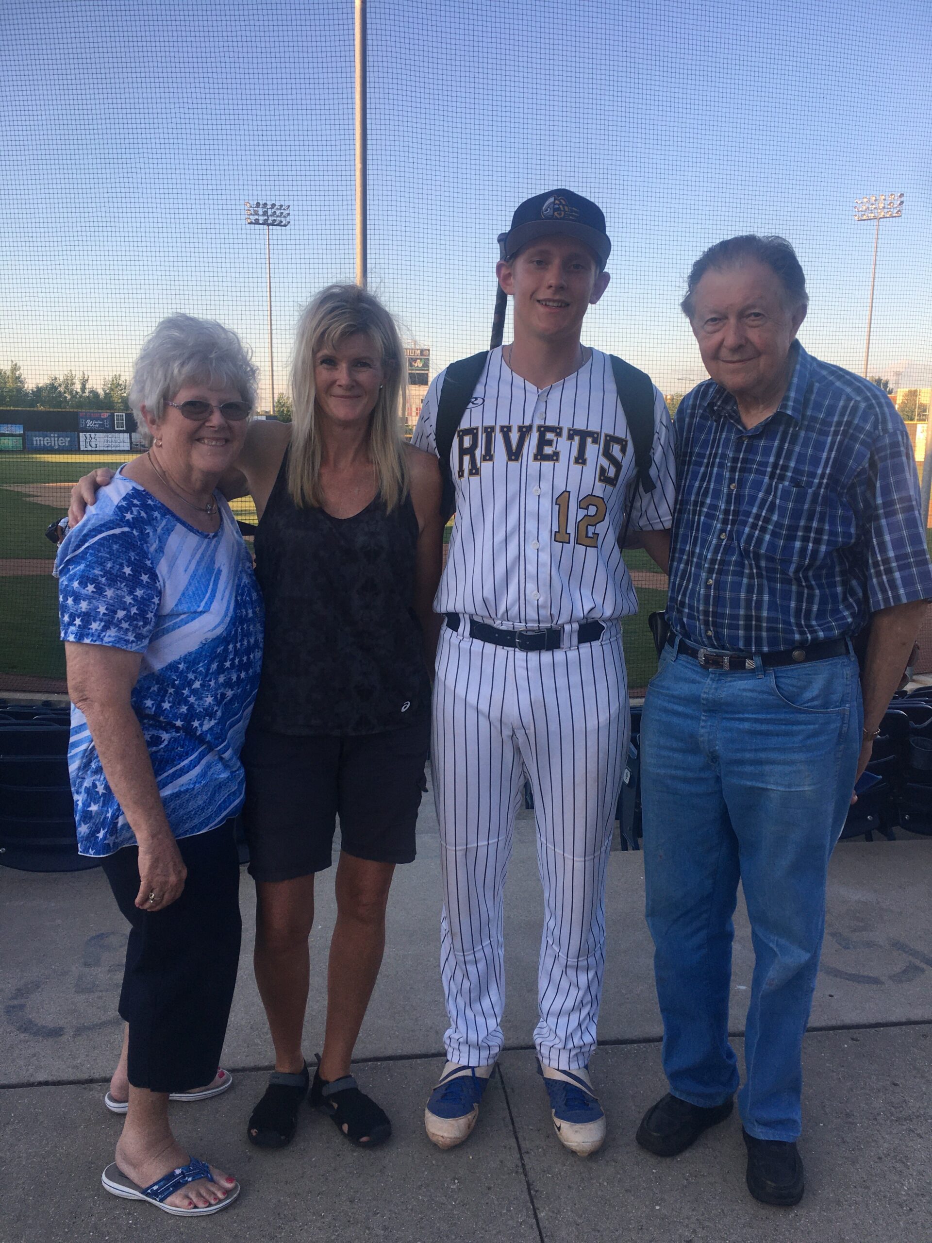 Group of four people posing at a baseball field; a smiling teen in a Rivets jersey (#12) stands with two women and an older man.