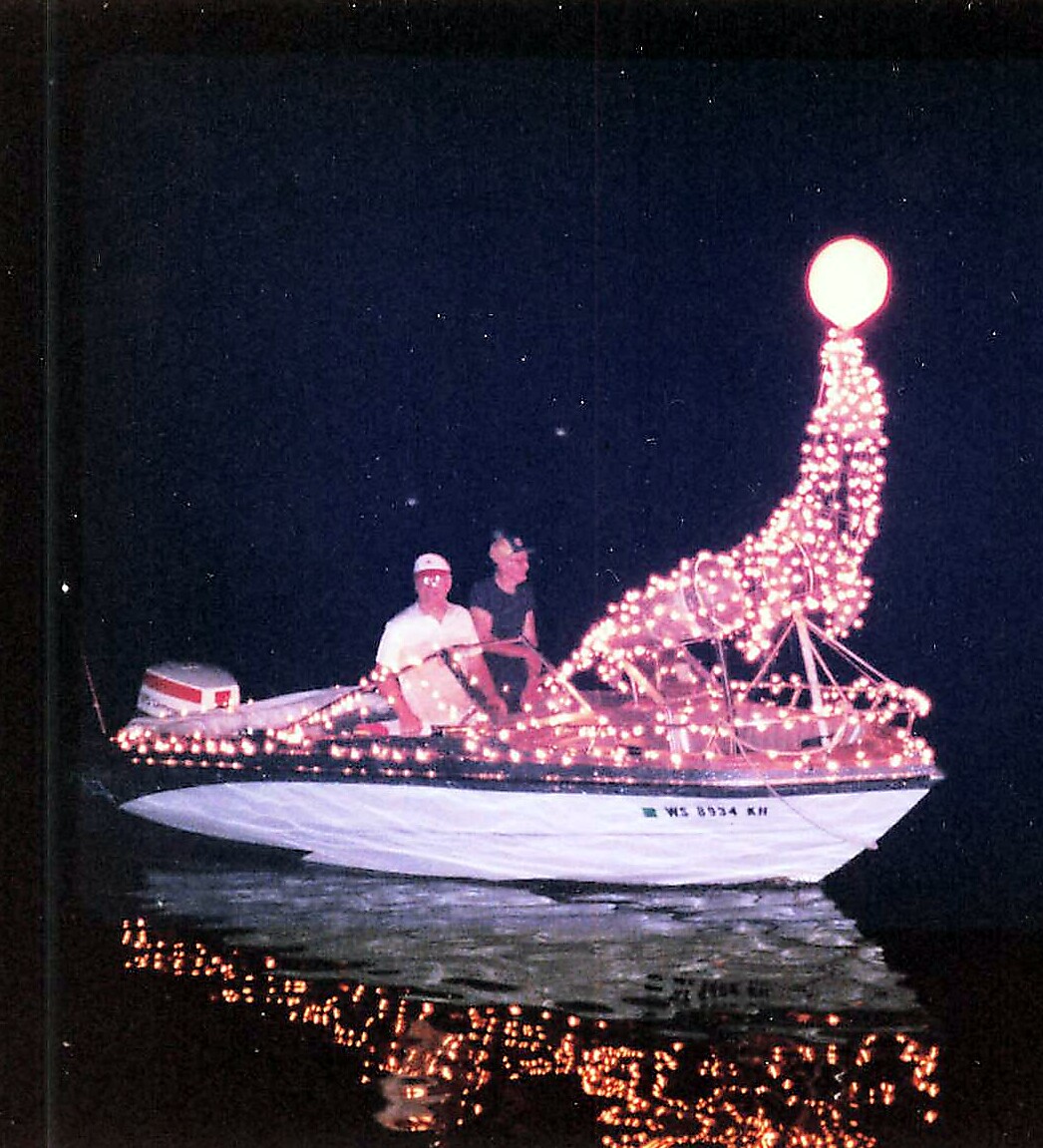 Boat at night wrapped in pink string lights forming a mermaid figure, with two passengers onboard.