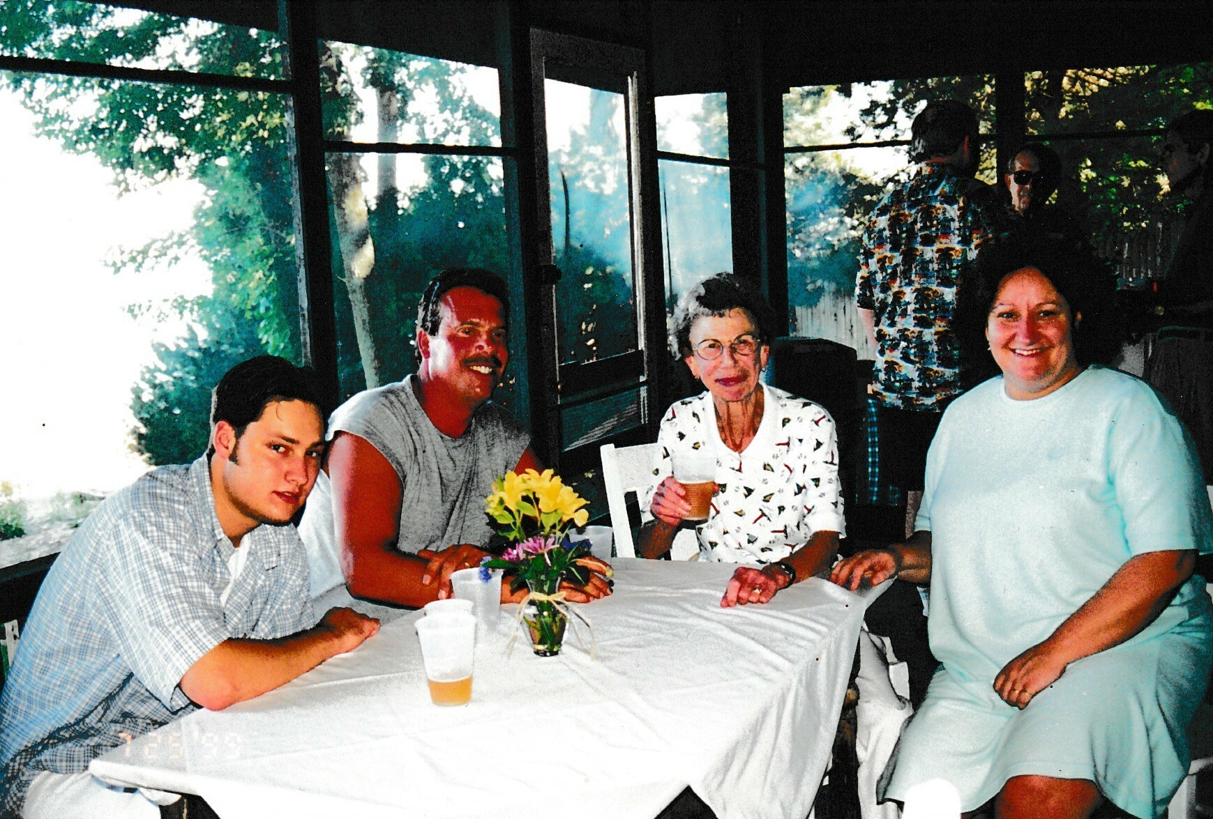 Four people sit around a table with a flower centerpiece, smiling at a casual outdoor gathering.