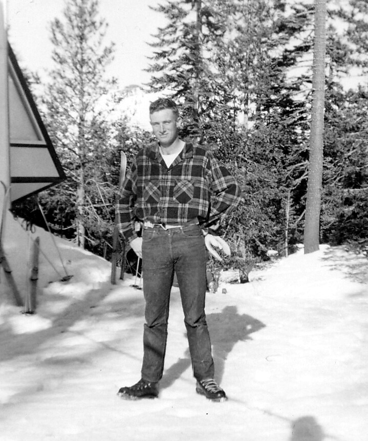 Man in a plaid shirt and jeans standing in snow with skis and trees in the background.