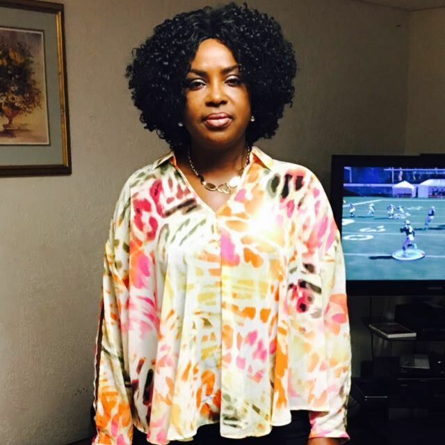 Woman with curly black hair wearing a bright abstract blouse, standing in a living room with a framed painting and a TV in the background.