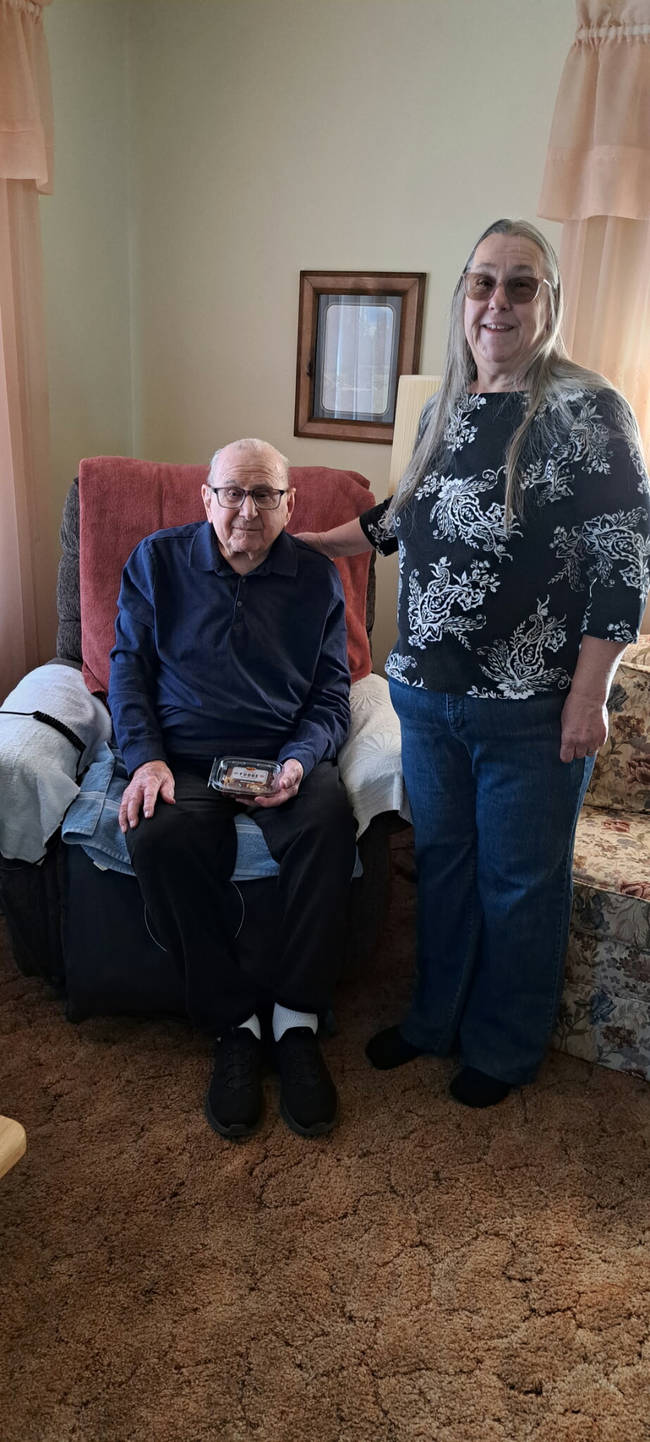 An elderly man seated in a chair holding a small plastic container, with a woman standing beside him in a living room.