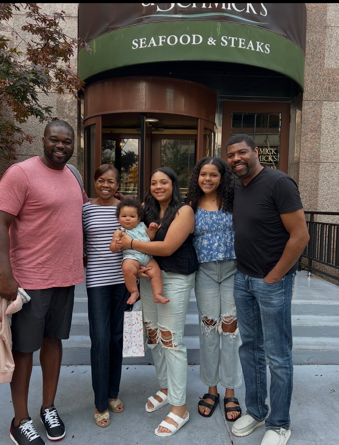 Family group posing together outside a restaurant with a green awning that reads 'SEAFOOD & STEAKS' and a revolving door behind them, smiling.