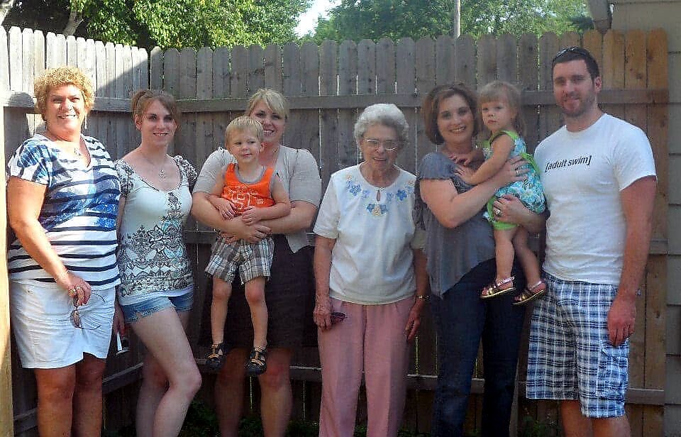 A multi-generational family group stands together in a backyard, smiling, with a wooden fence behind them.