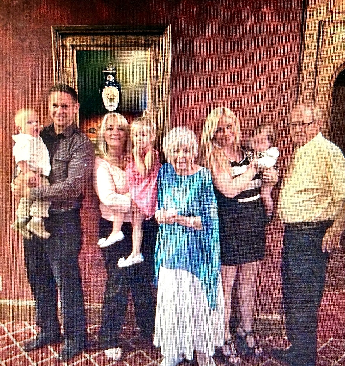 A multigenerational family portrait: grandparents, adults, and several small children posing together in a living room with a red wall and framed painting behind them, two babies held by adults.