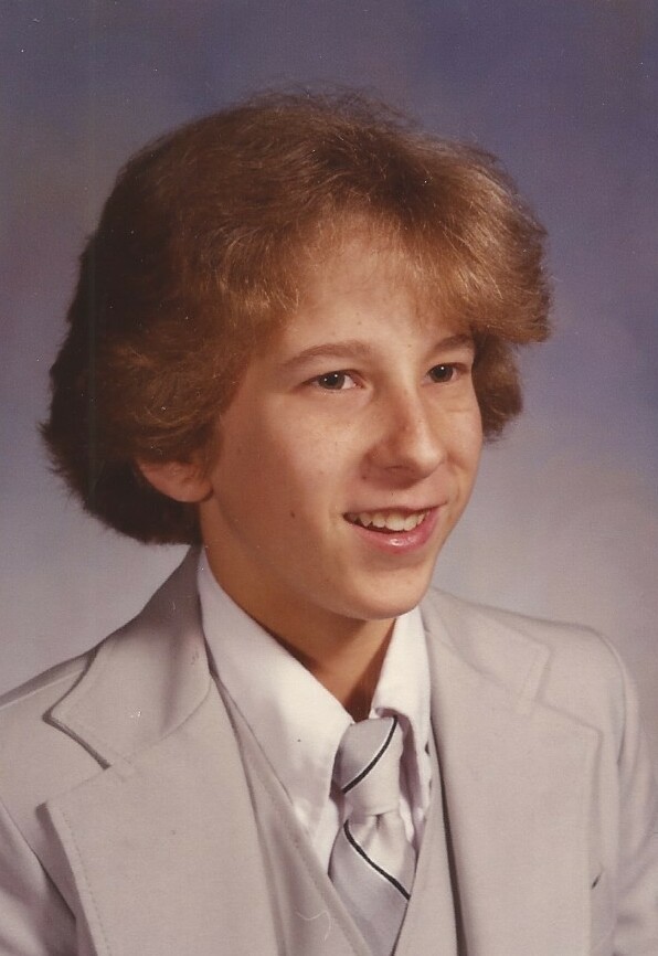 Young boy in a light gray suit and tie posing for a school portrait, smiling at the camera.