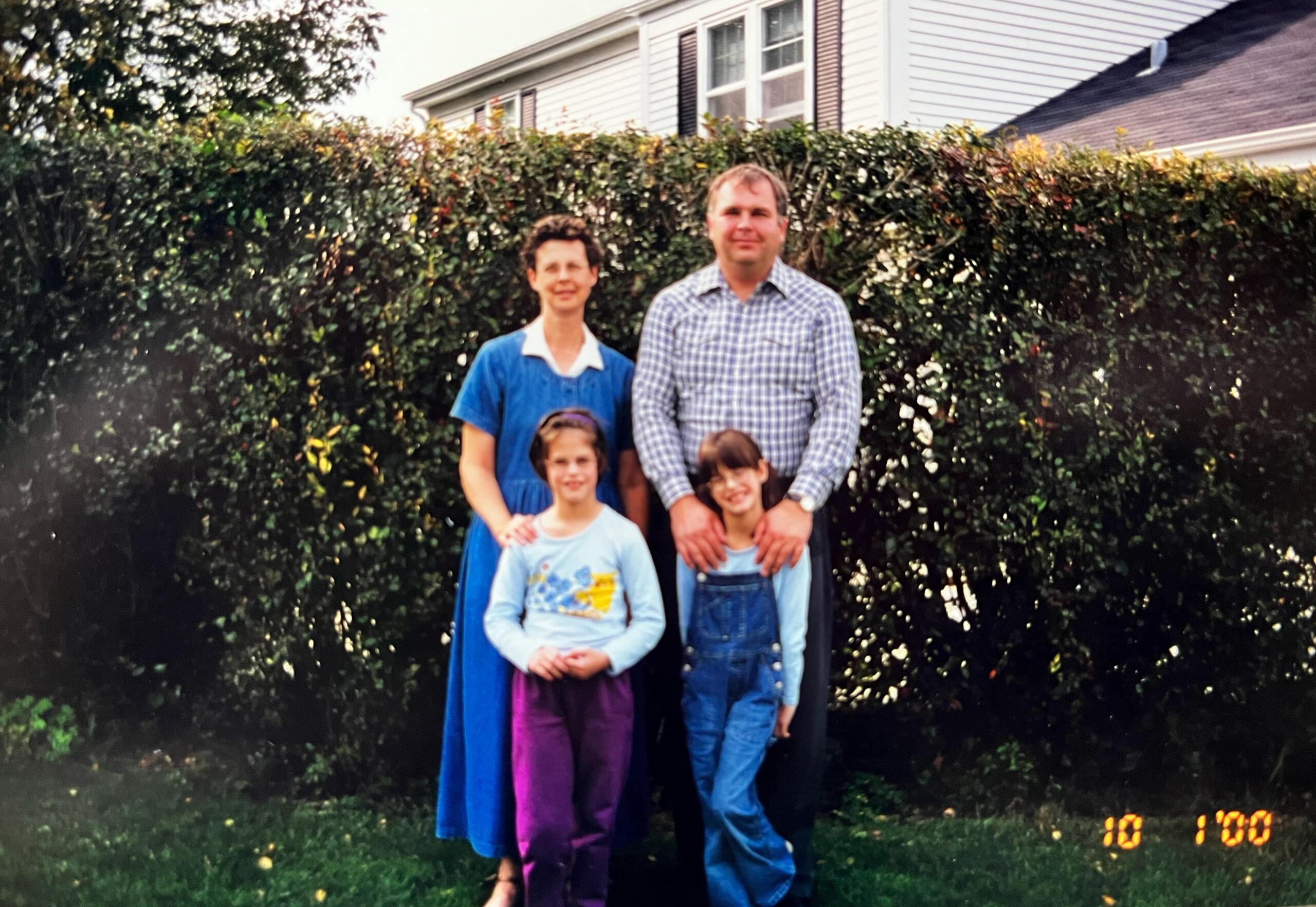 Family of four posing outdoors in front of a tall hedge with a suburban house behind them. Two adults with two children.