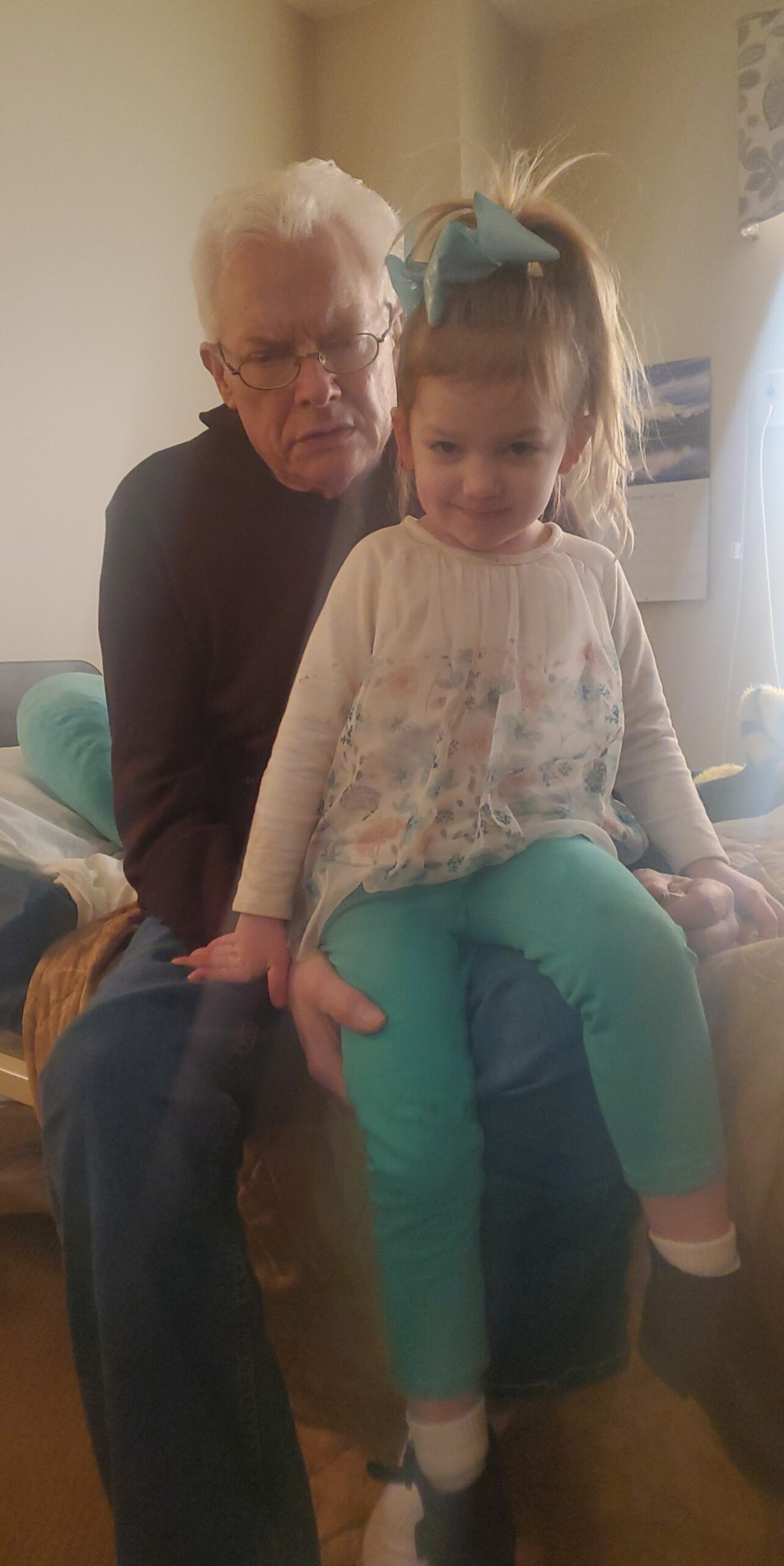 Grandfather sitting on a bed with a young girl on his lap, both looking at the camera; girl wears teal leggings and a large teal bow in her hair.