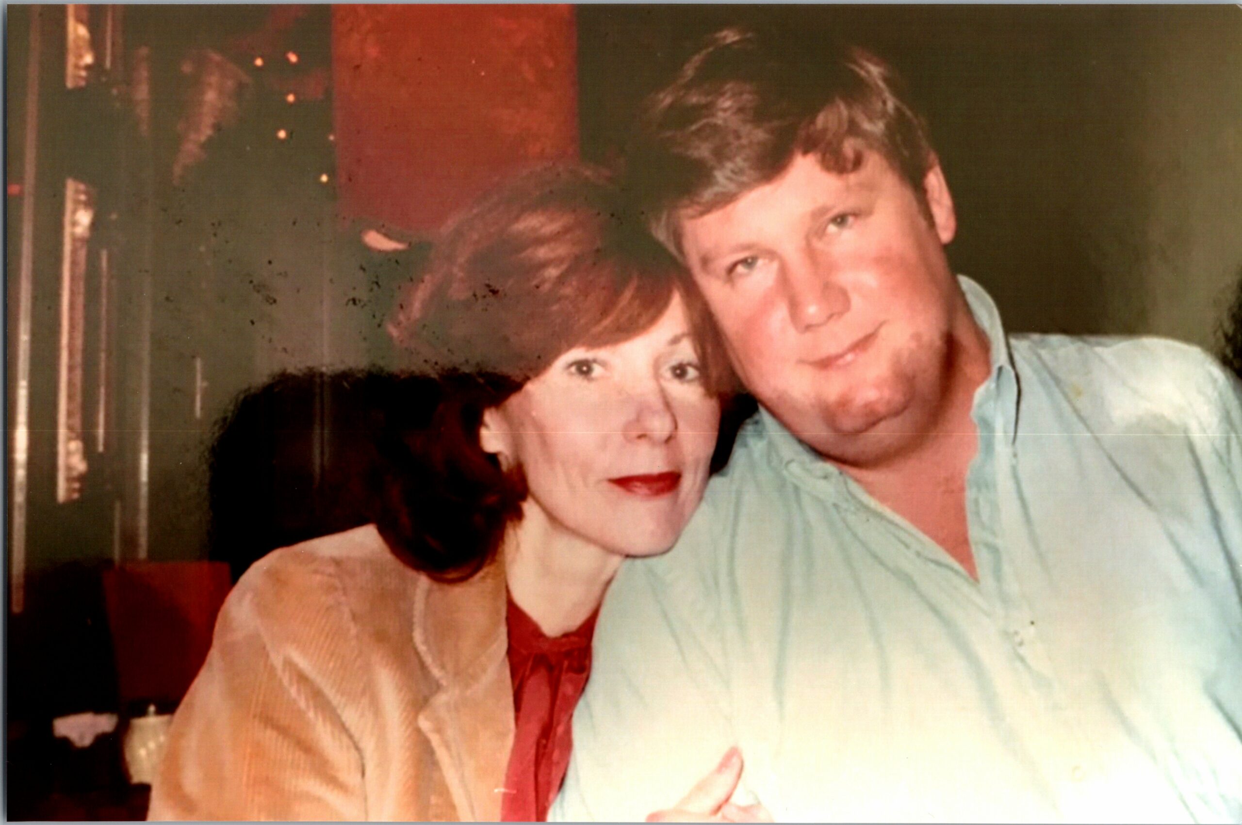 Couple posing closely for a photo: woman with short dark hair and red lipstick, man with light brown hair in a pale blue shirt.