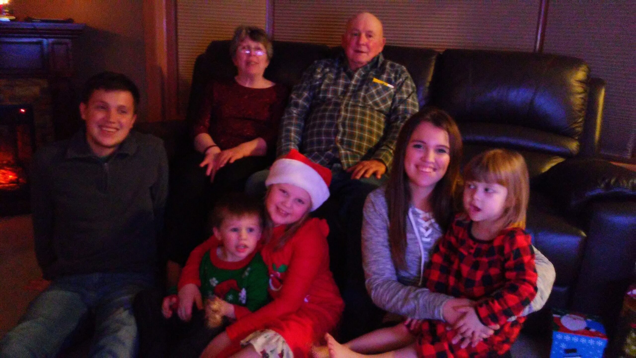 Family group sitting on a living room sofa for a holiday photo, kids in festive outfits and Santa hat smiling at the camera. Note: 7 people present across sofa and floor, warm family scene.