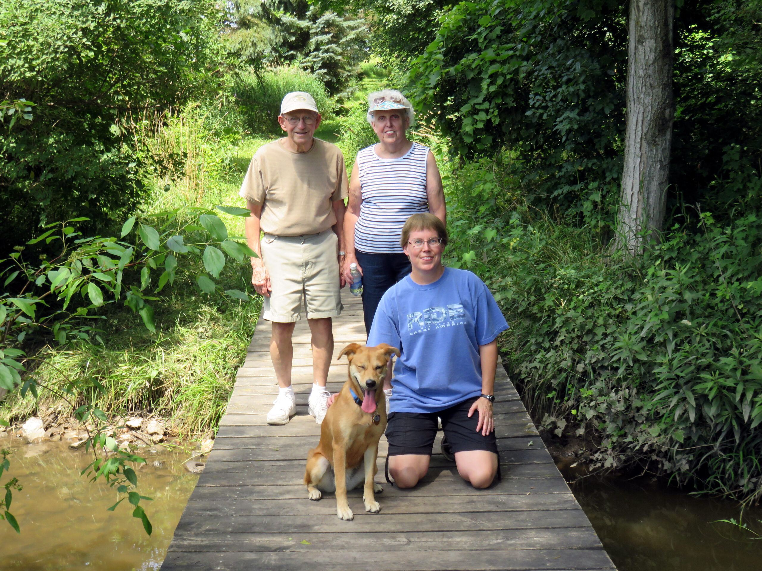 Three adults and a tan dog posing on a wooden boardwalk surrounded by green vegetation near a muddy pond.