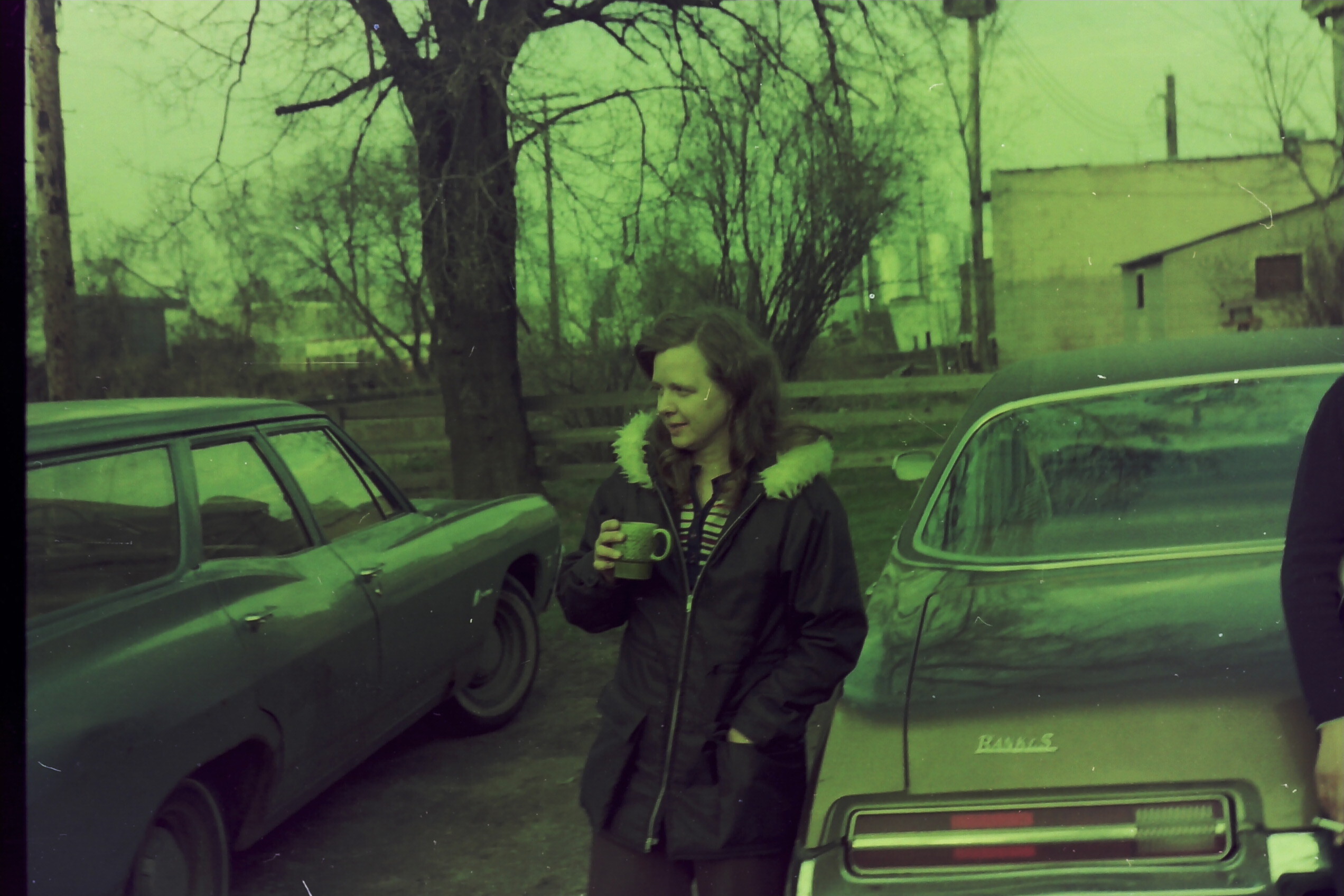 Woman in a dark coat with a fur-trimmed hood holds a mug while standing between two parked cars in a yard with trees in the background.