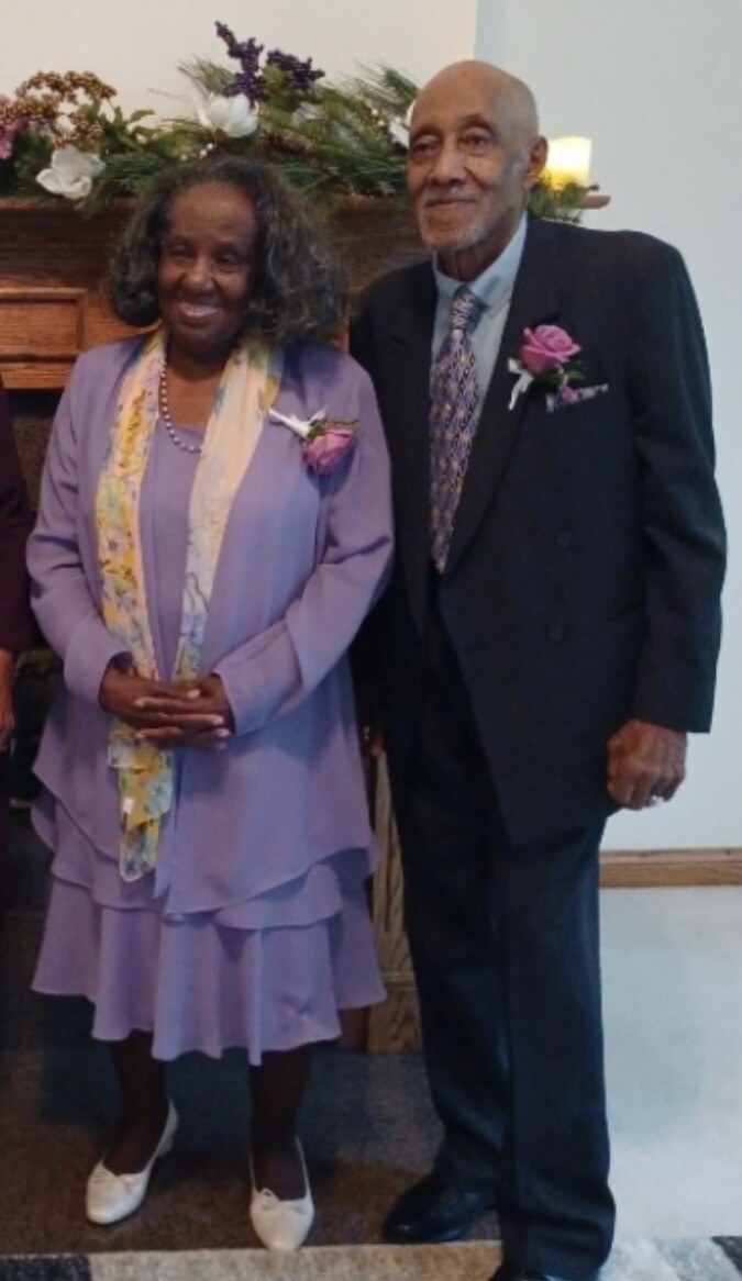An elderly couple standing together at a formal event, woman in a lavender dress and scarf, man in a dark suit with a purple boutonniere.