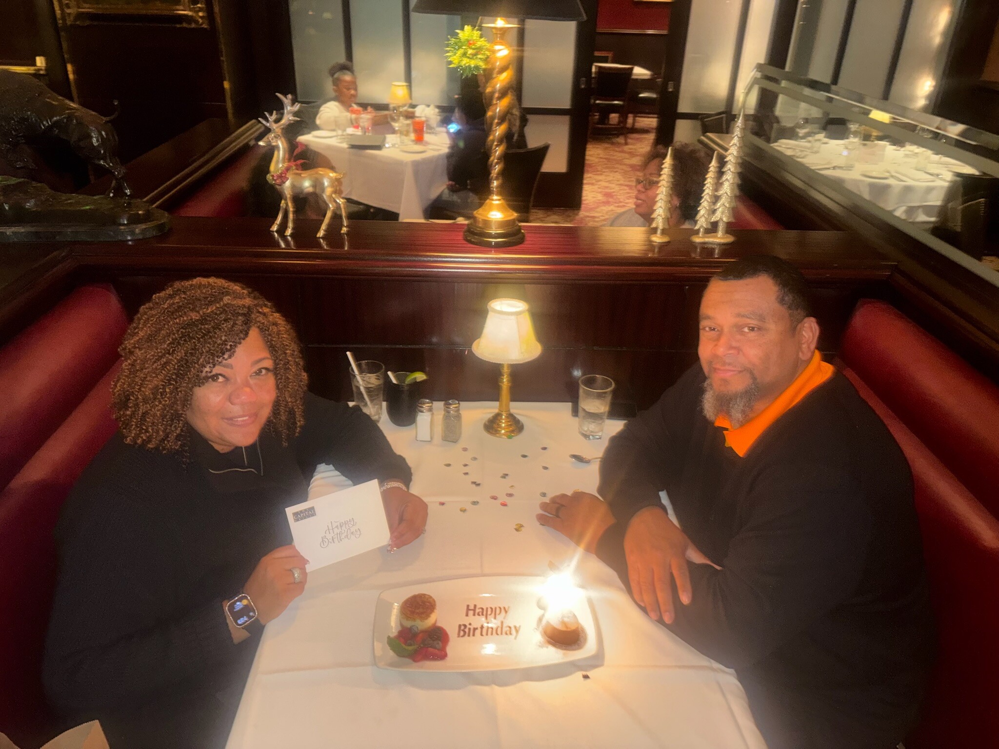 Two people sit across from each other at a restaurant table, celebrating a birthday with a plate that says ‘Happy Birthday’ and a card on the table nearby.