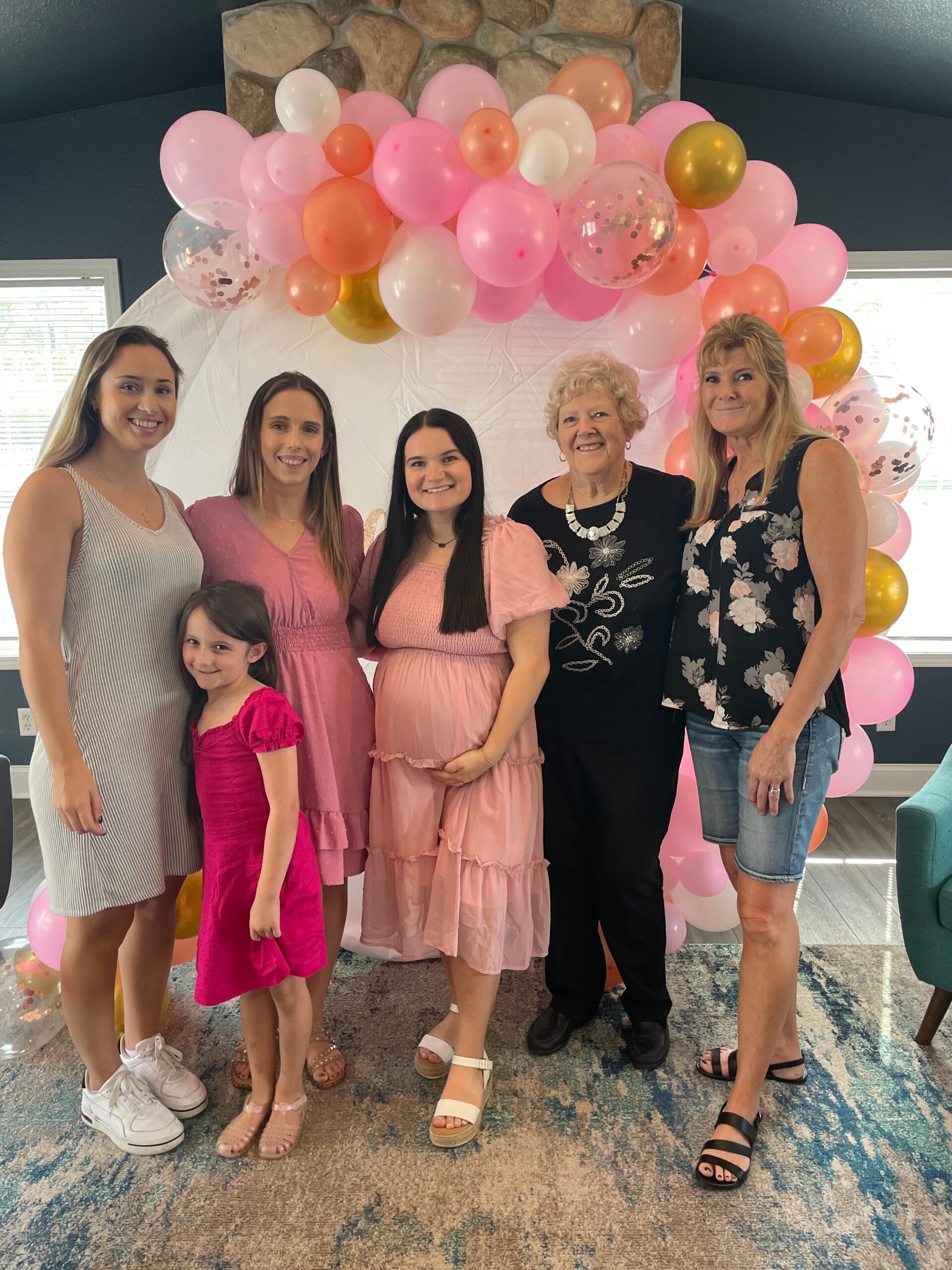 Six women and girls pose together in front of a pink balloon arch at a celebration.