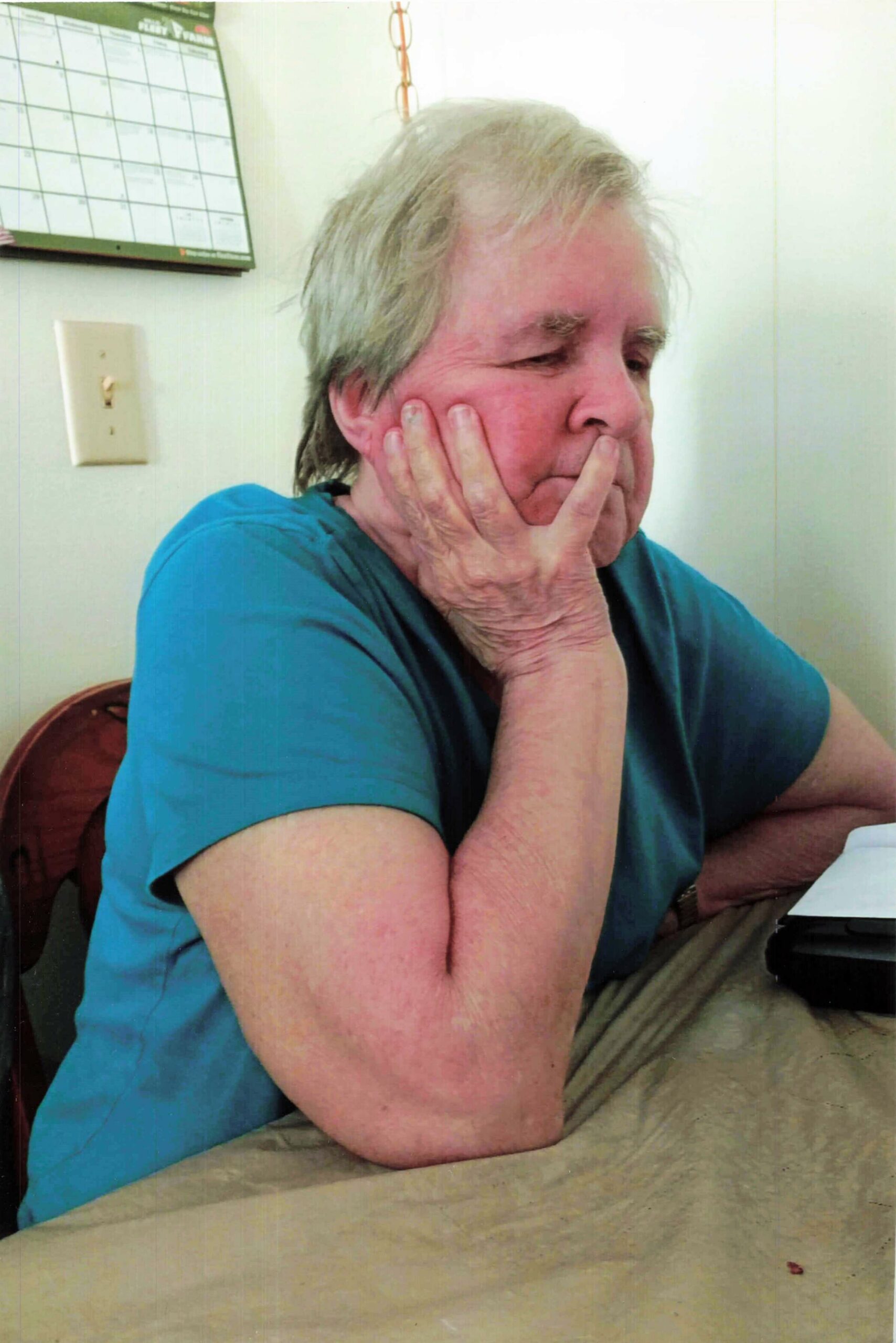 Older woman with short gray hair in a teal shirt rests her chin on her hands, looking down pensively indoors with a calendar on the wall behind her.