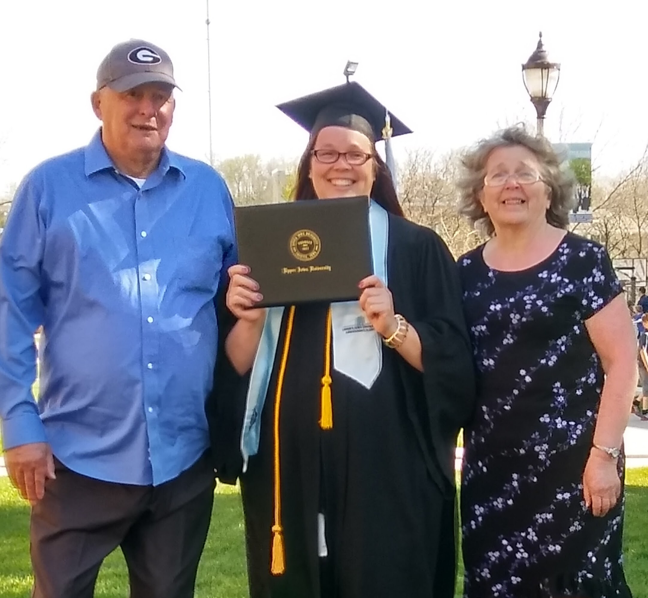 Three people pose outdoors for a graduation photo: a graduate in cap and gown holds a diploma, flanked by a man in a blue shirt and a woman in a floral dress.