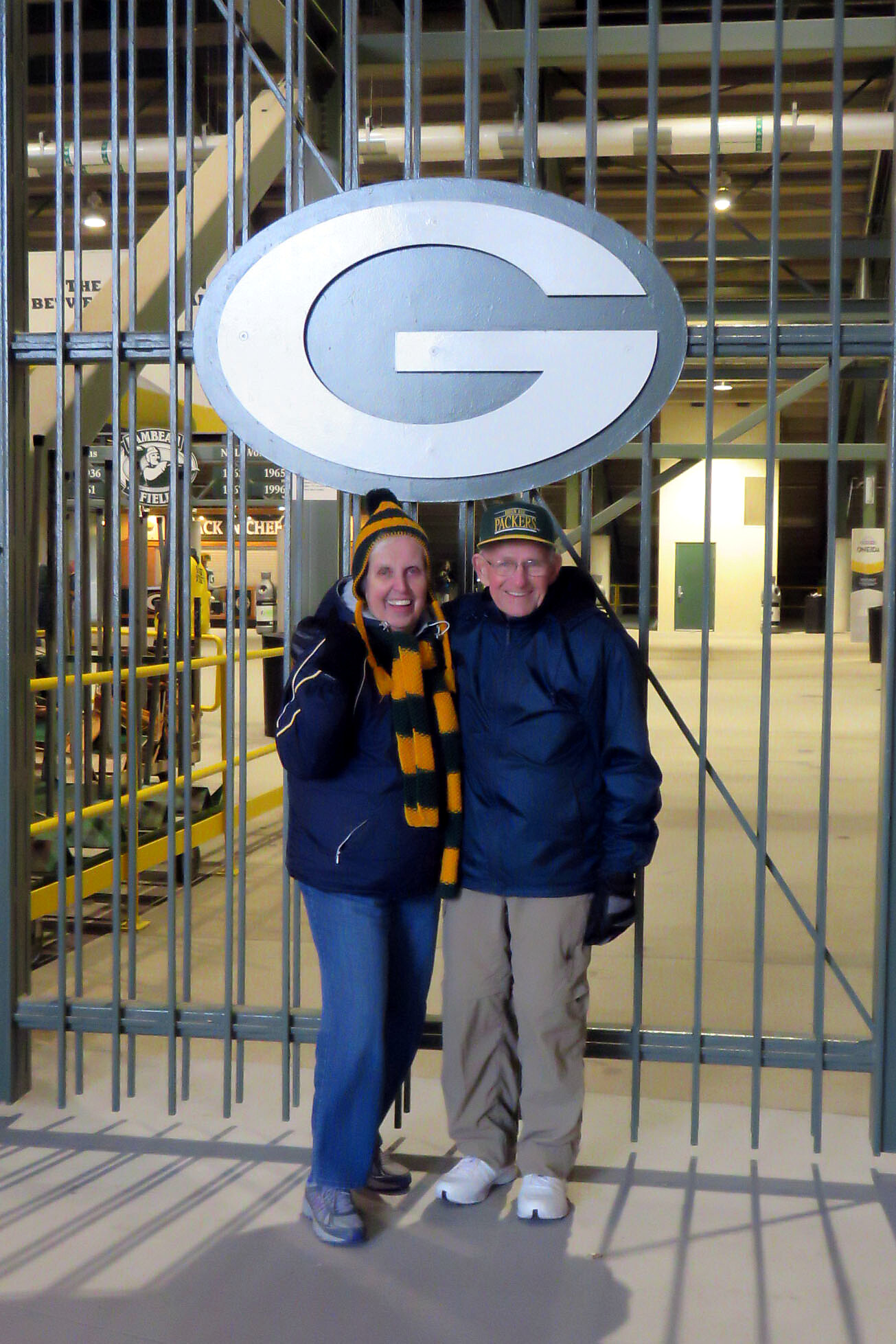 Smiling couple in winter coats pose behind a large oval green 'G' logo on a gate inside a sports stadium railing area.