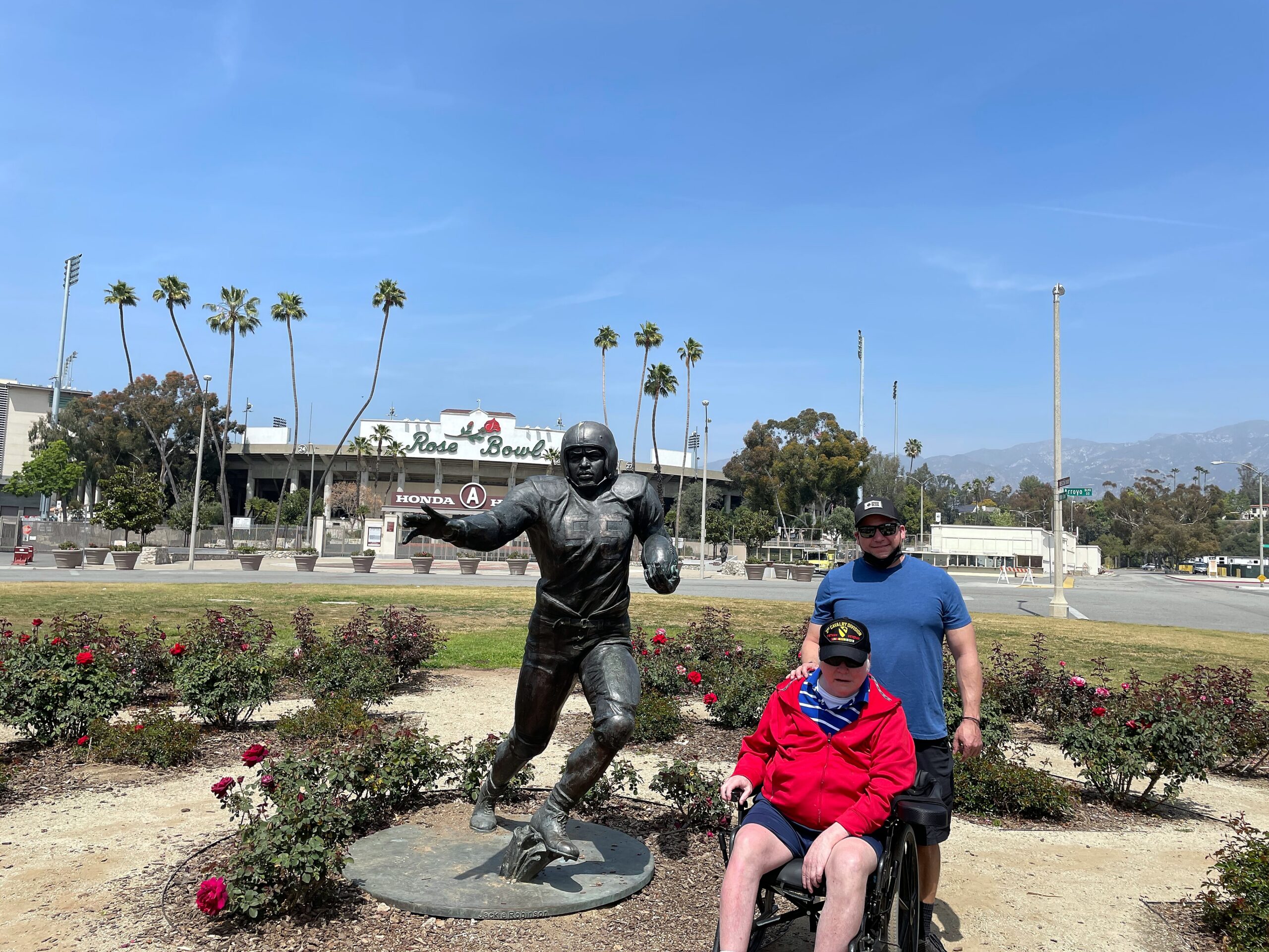 Bronze statue of a football player in a garden in front of the Rose Bowl stadium, with a man in a wheelchair and a boy in blue nearby among rose bushes and palm trees.