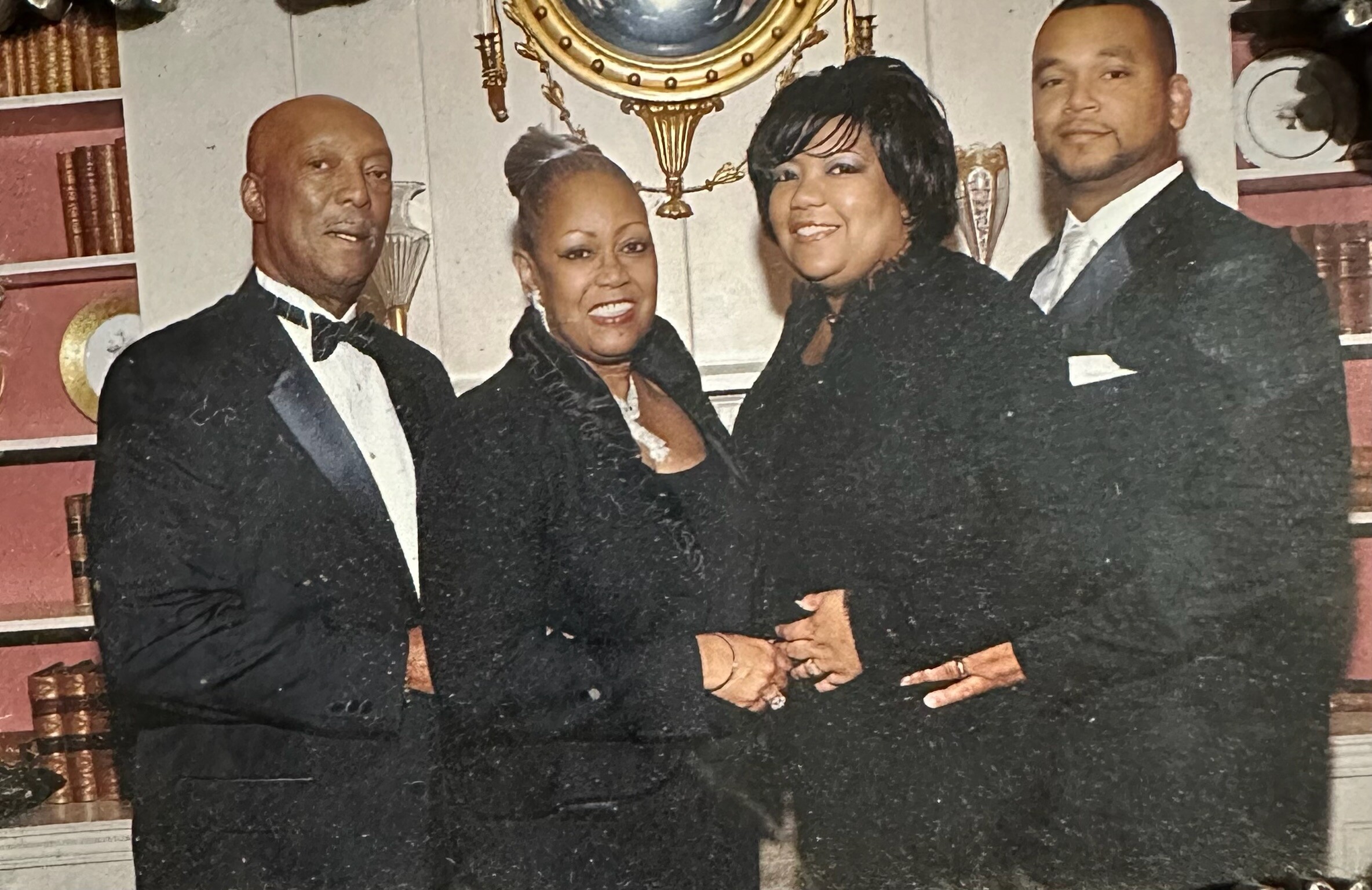 Four adults in formal black-tie attire posing together in a home setting with a bookshelf and decorative items in the background.