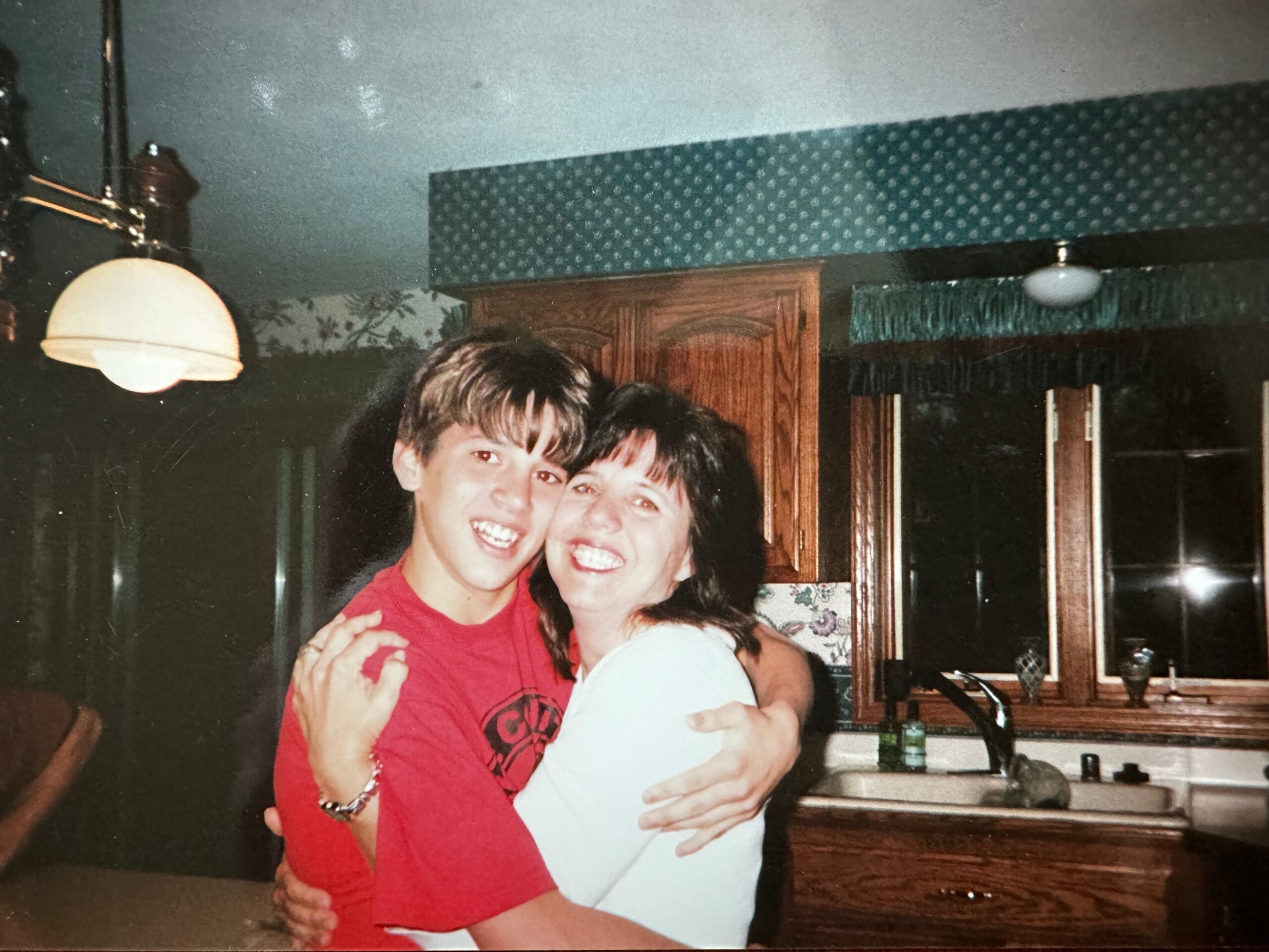 Two people hug and smile at the camera in a home kitchen with dark cabinets in the background.