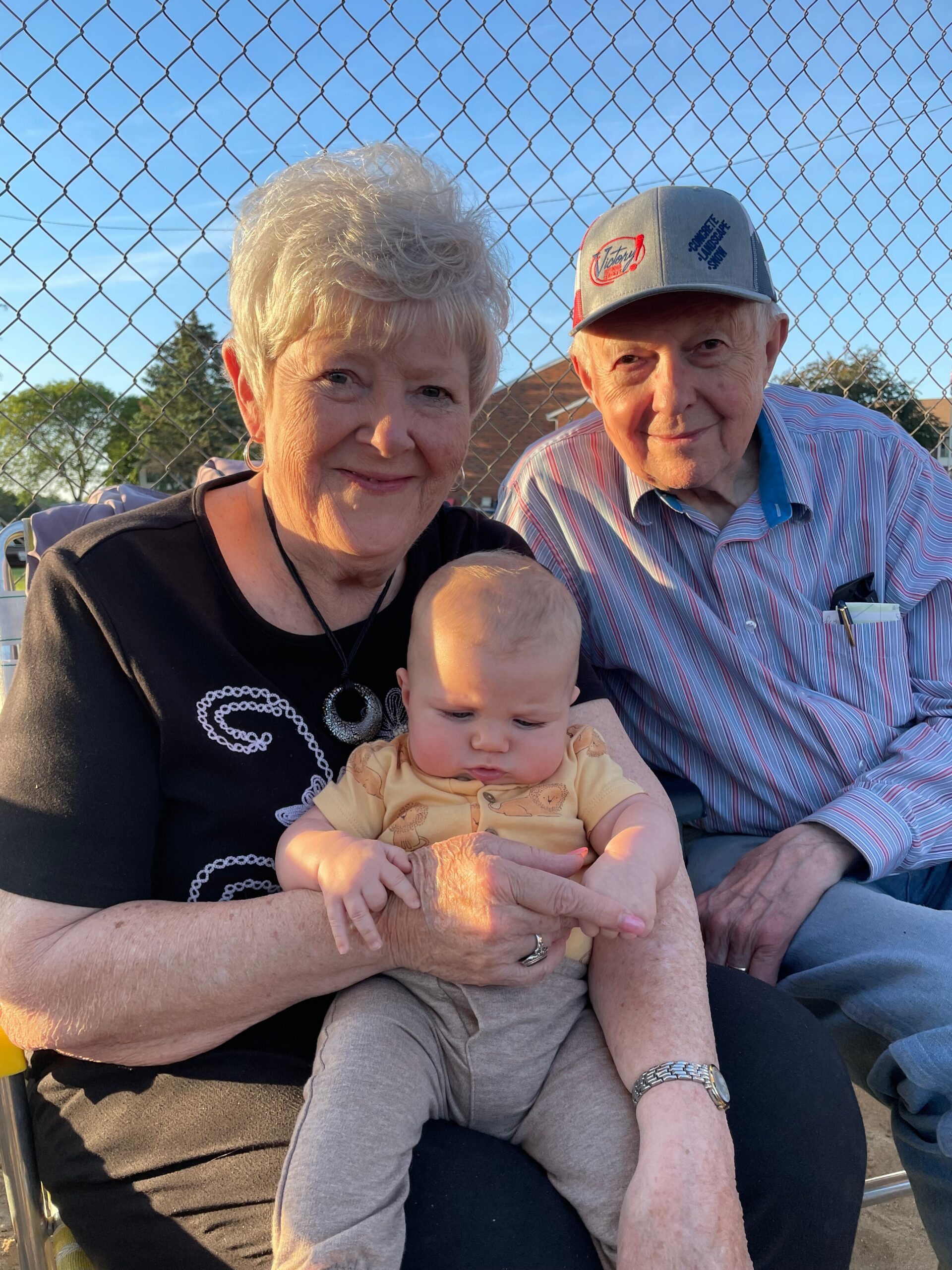 Grandparents sit with a baby between them, behind a chain-link fence on a sunny day.