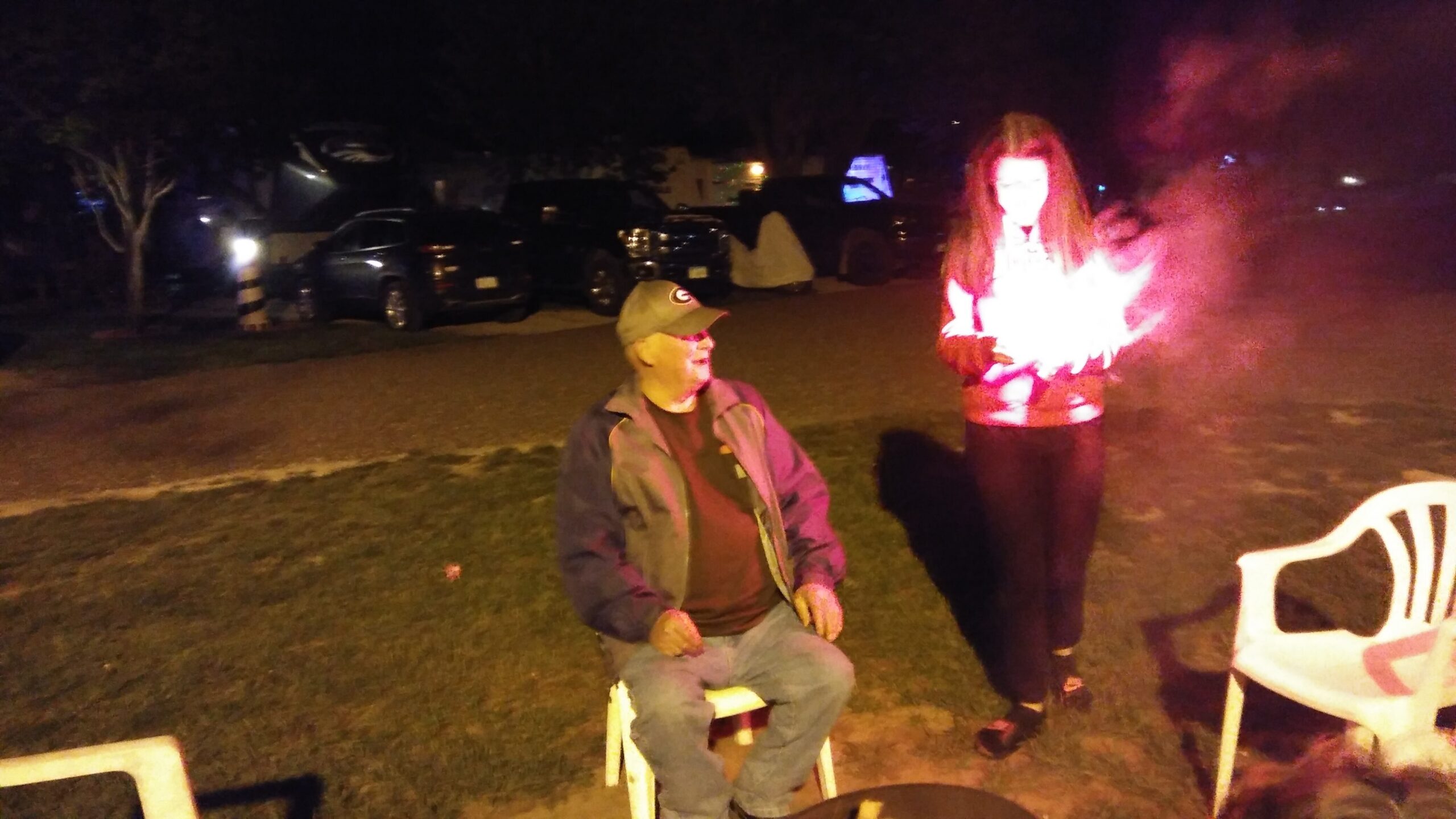 Nighttime outdoor gathering: man in a cap and jacket sits on a white chair, smiling, while a person in a red jacket stands nearby with a bright flame or light source.