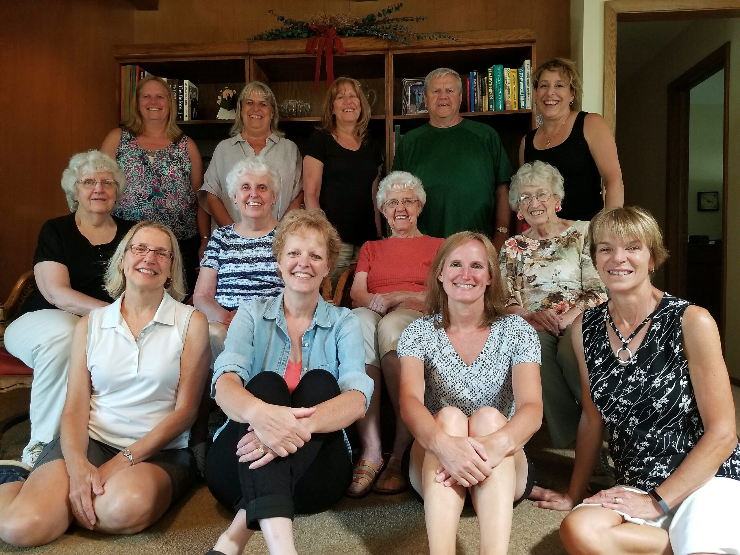 Group of smiling adults posing for a photo in a living room with a bookcase behind them. A mix of men and women, several seated in front row and others standing in back row.