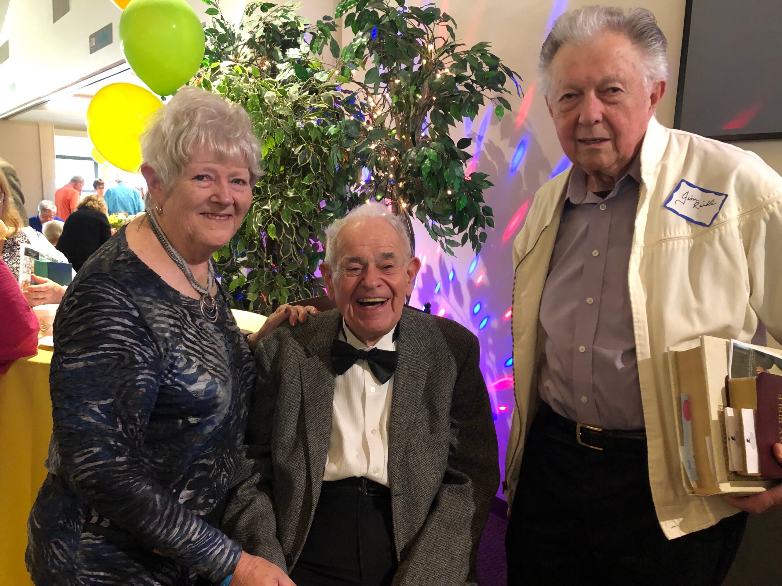 Three elderly friends posing for a group photo at a party, with balloons and a lit plant in the background.