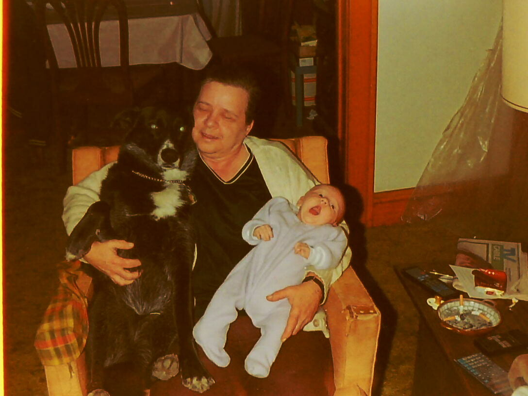 Older woman sits in a chair, holding a black dog on one side and a baby in a blue onesie on her lap in a cozy living room.