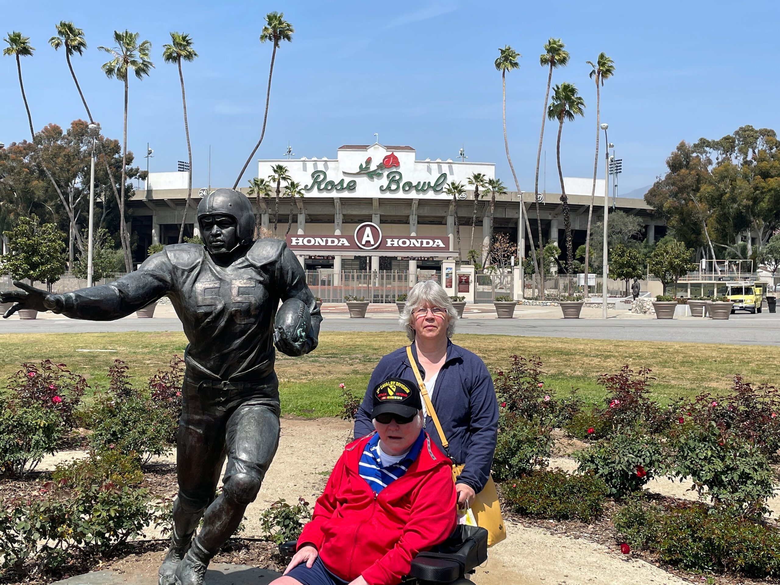 Bronze football player statue reaching forward in front of the Rose Bowl stadium, with visitors and tall palm trees in the background.