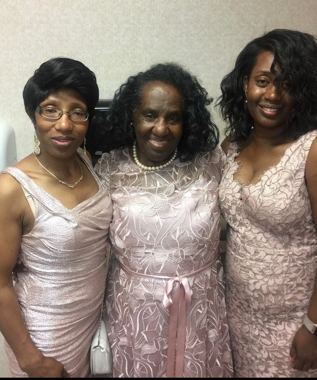 Three women in pink lace dresses posing together and smiling at the camera with a neutral backdrop.