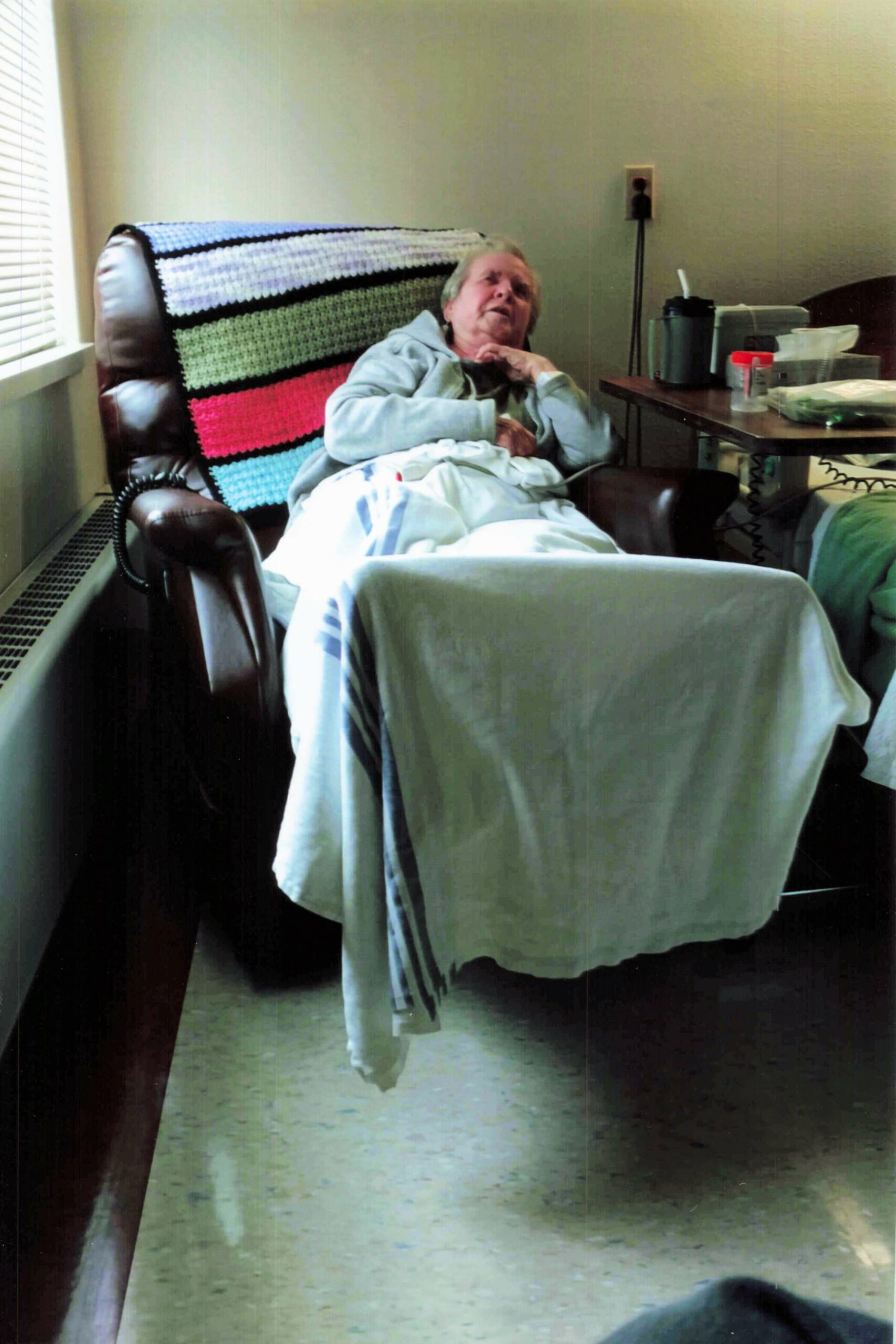 Elderly person resting in a recliner, covered with a colorful striped crocheted blanket, hands folded on the chest in a room with a table of medical supplies nearby.