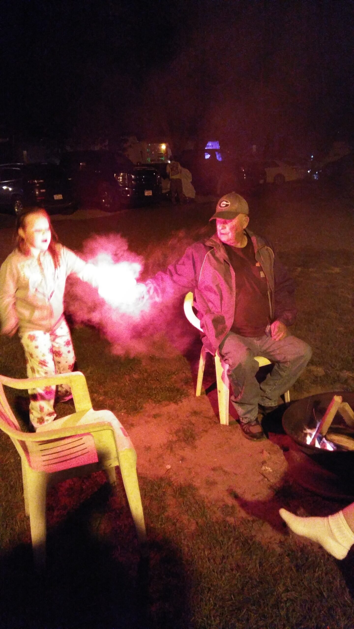 Nighttime backyard scene with a man sitting on a chair by a campfire and a girl releasing pink smoke.