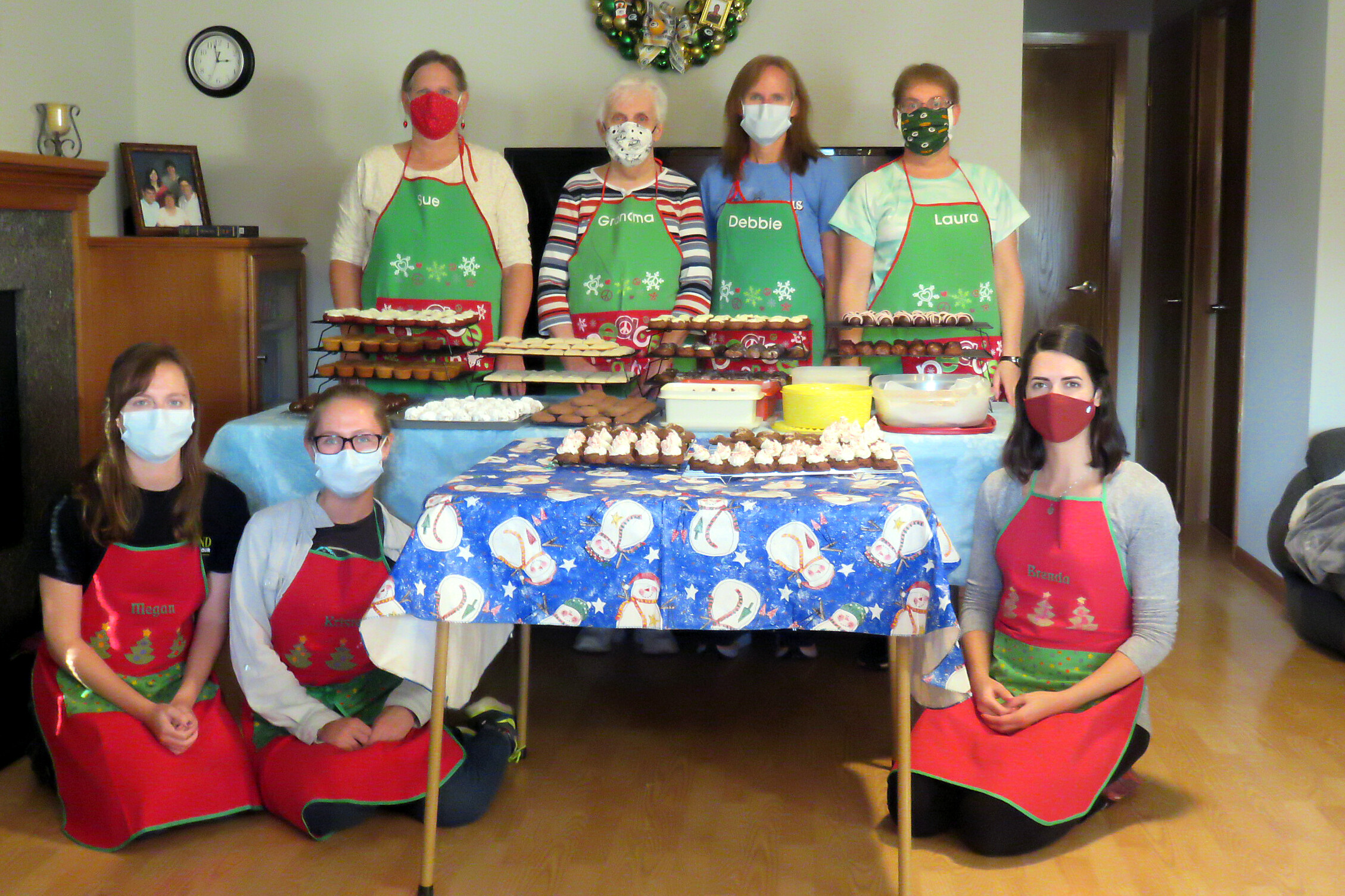 Group of people in festive red-green aprons and face masks posing behind a table of cookies and baked treats for a holiday bake sale or gathering.