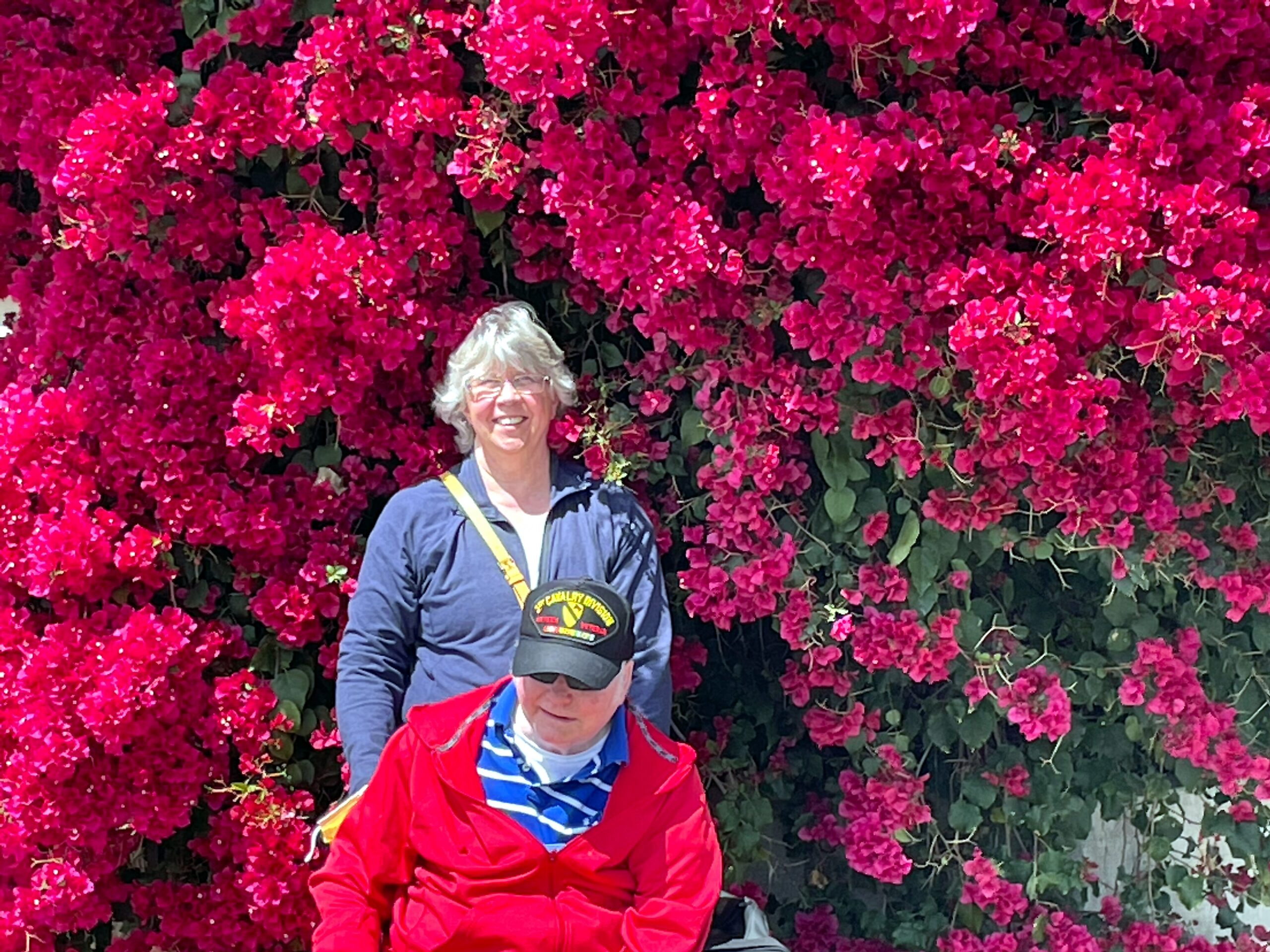Two people posing in front of a wall of bright pink bougainvillea blooms, smiling at camera.