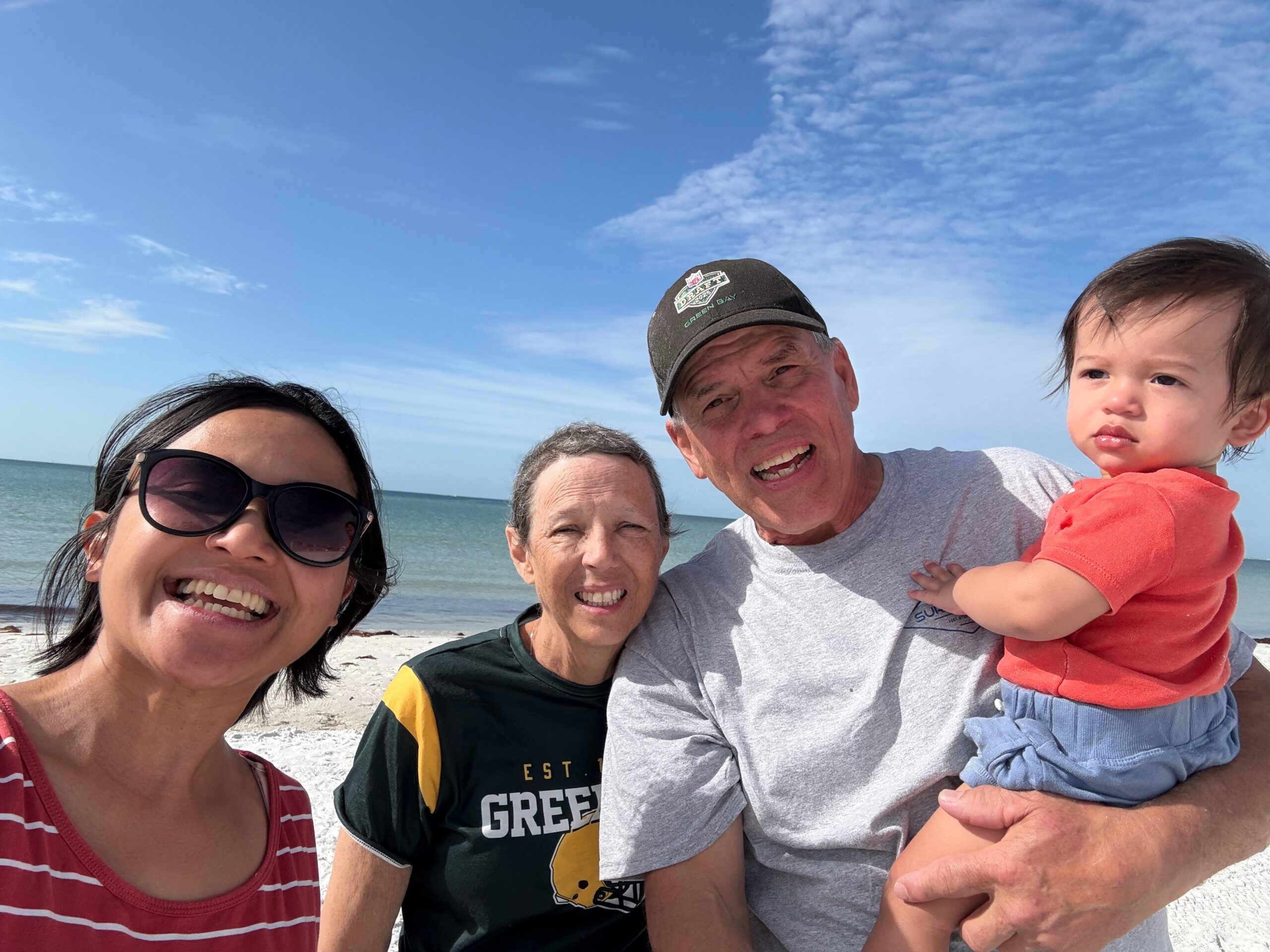 A smiling family selfie on the beach: woman with sunglasses, man in a gray t‑shirt, and a toddler in an orange shirt held by another adult, with the ocean and blue sky behind them.