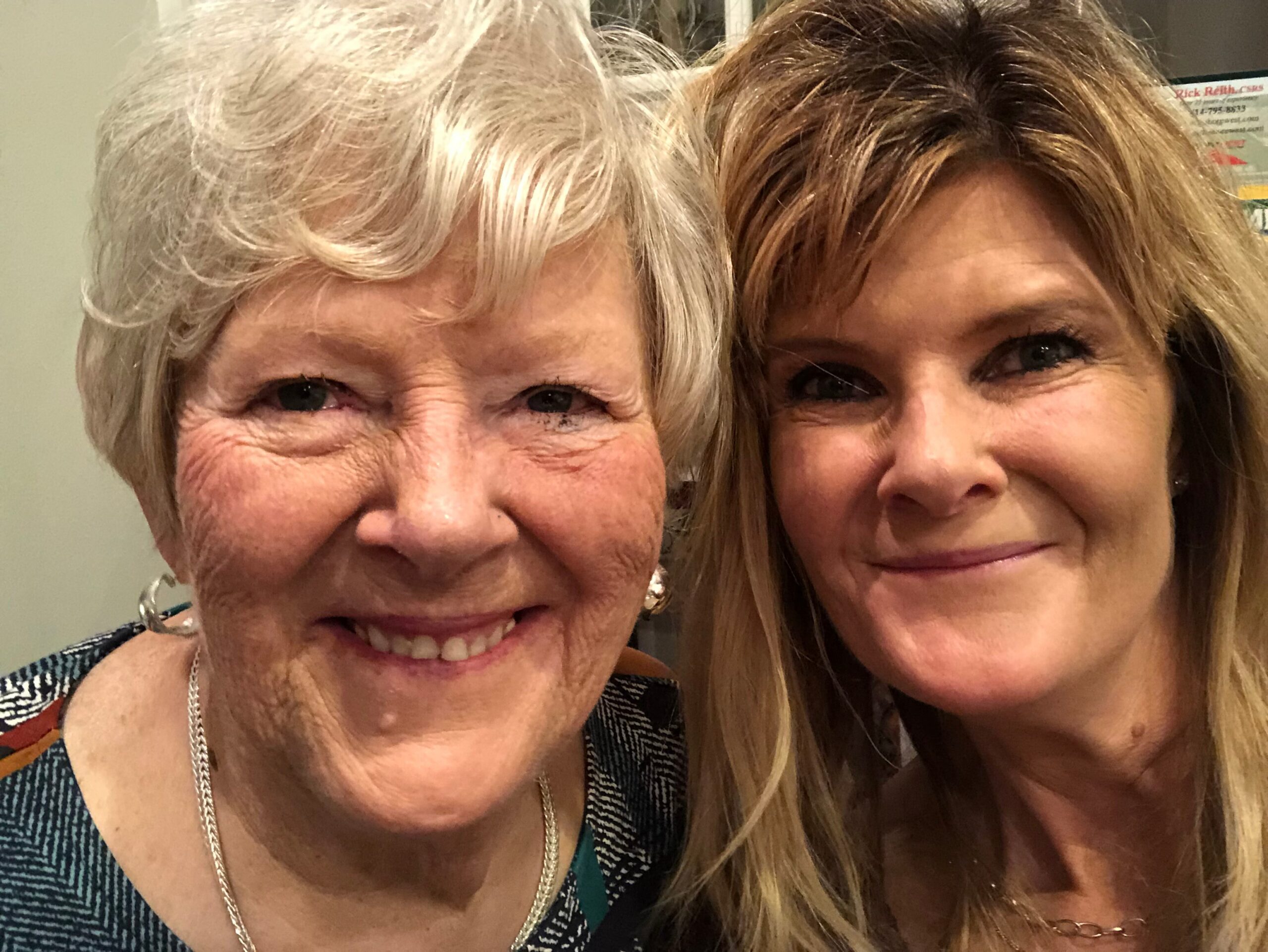 Two women smiling in a close-up selfie, showing their faces from the nose up with blond and light brown hair.