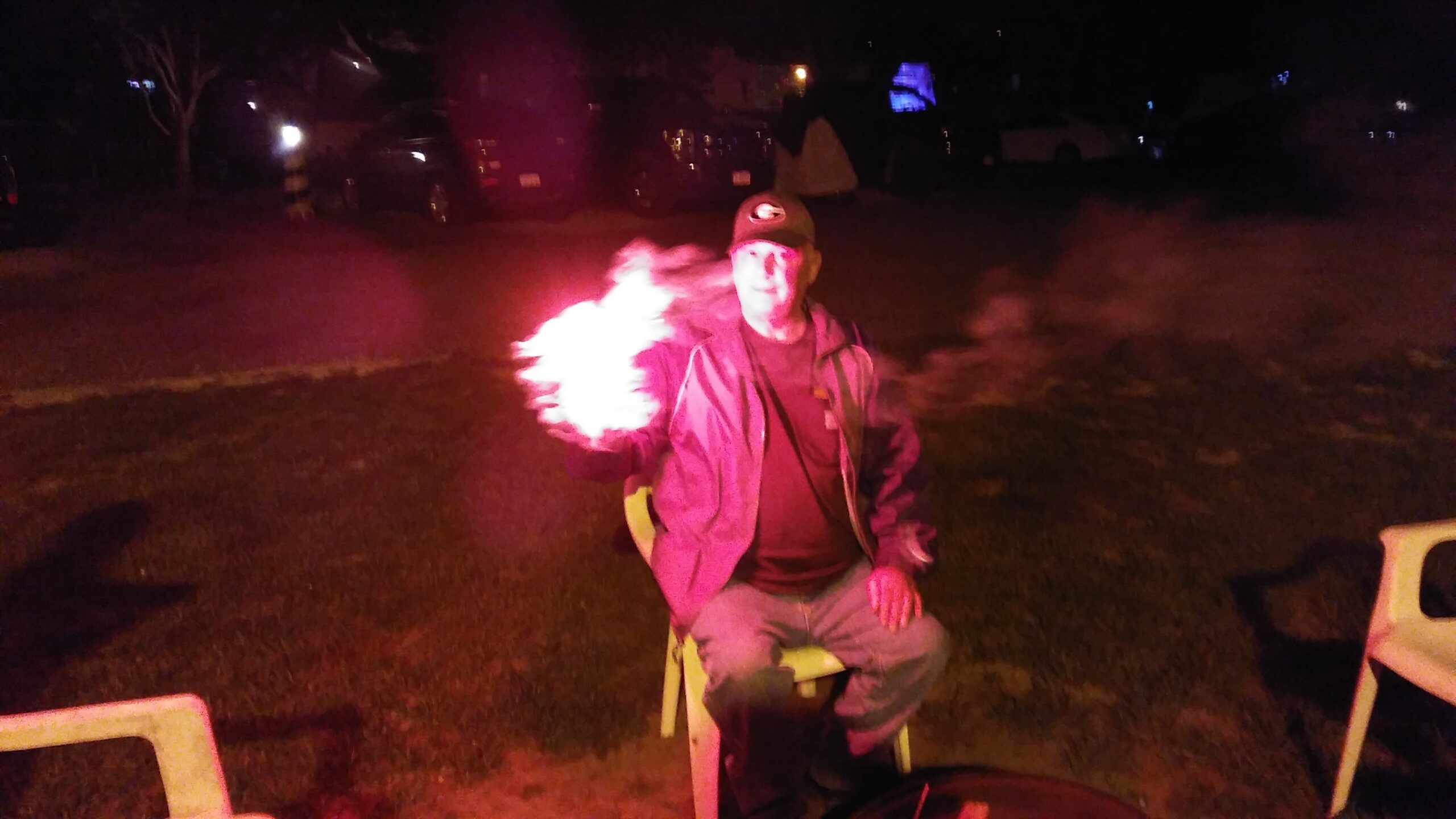 Man sitting on a white chair at night, holding a bright flame in his right hand beside his body.
