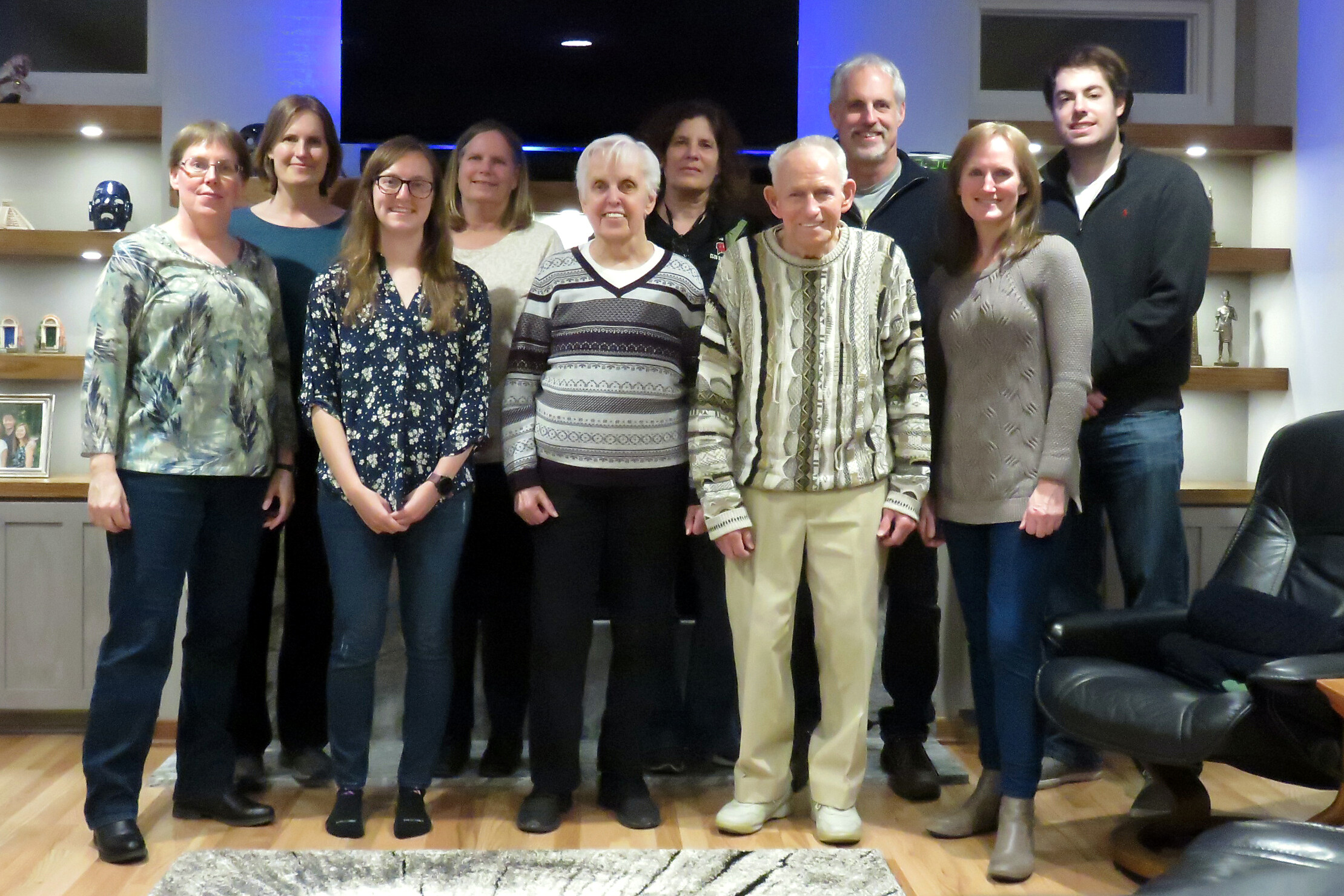 Group of eleven adults posing for a photo indoors, standing shoulder to shoulder in a room with shelves and a sofa visible behind them.