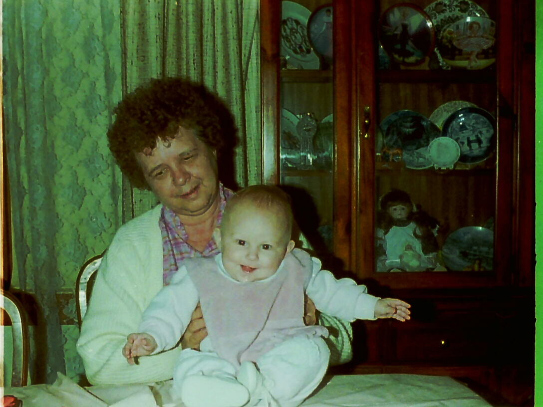 Man with curly hair holding a smiling baby on a dining table in a home setting. They sit near a wooden cabinet with plates and dolls inside.
